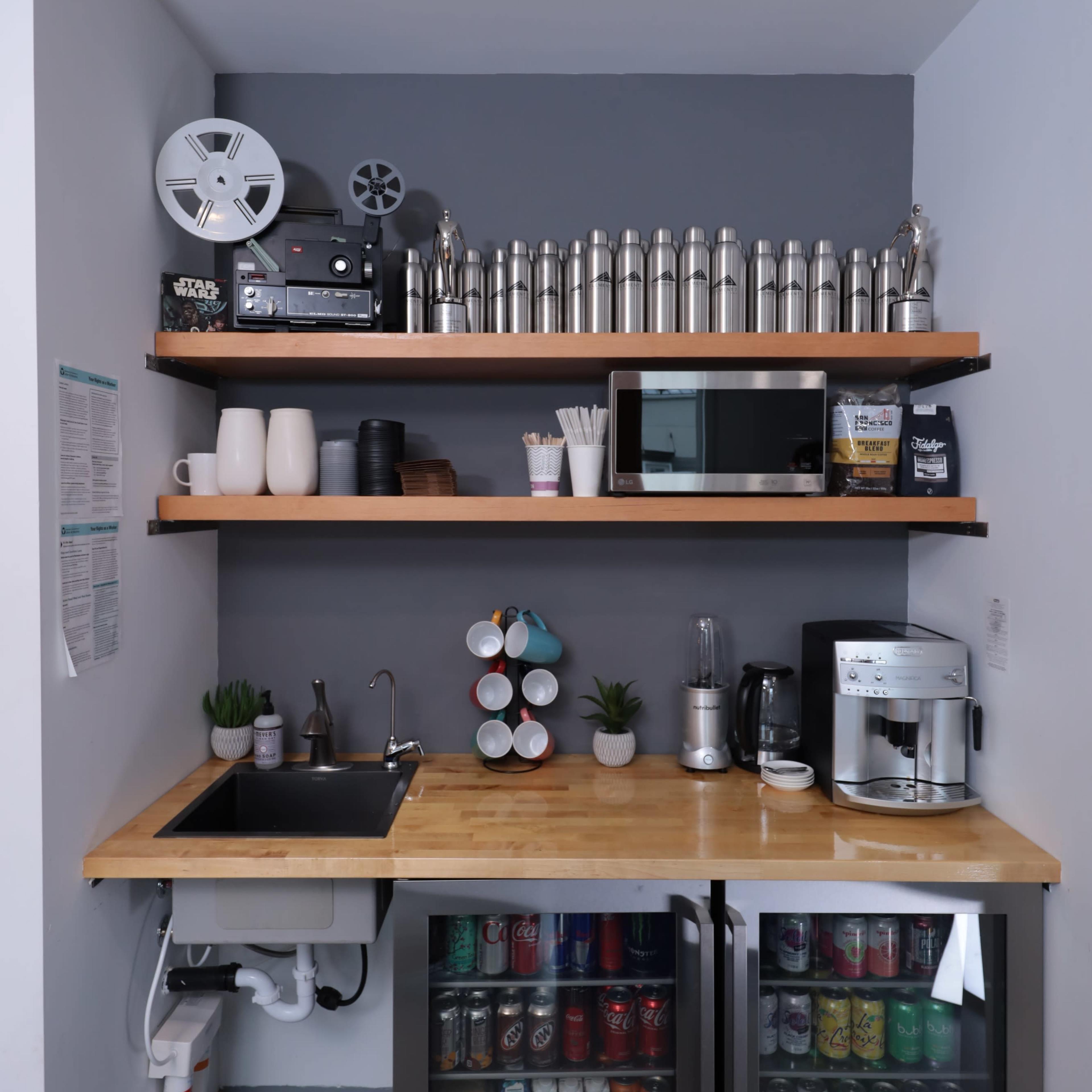 A compact kitchenette with shelves containing drink bottles, a coffee maker, a sink, and refrigerators stocked with beverages.