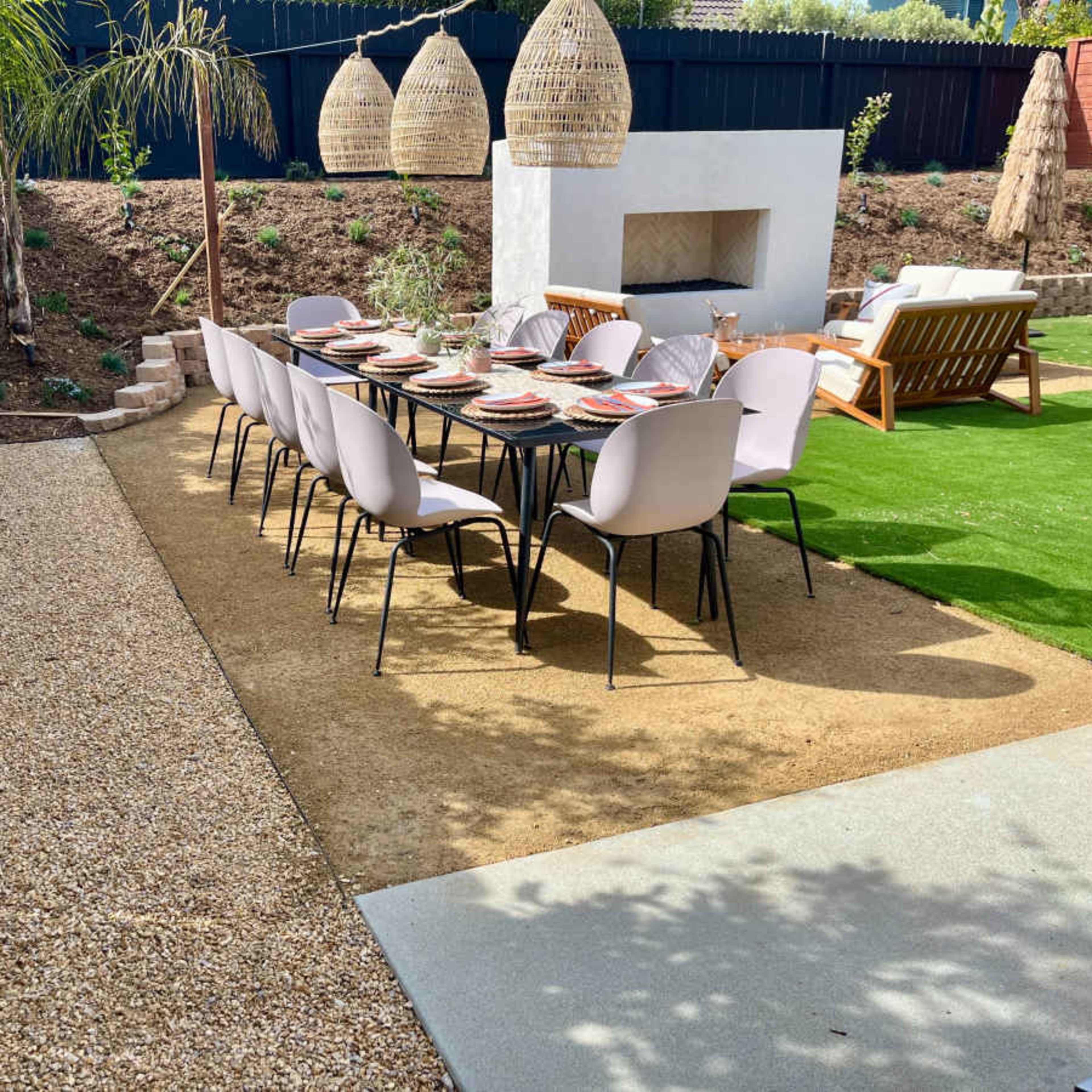 An outdoor dining area with a long table set for a meal, surrounded by a gravel path, a grassy area, and decorative hanging lights.