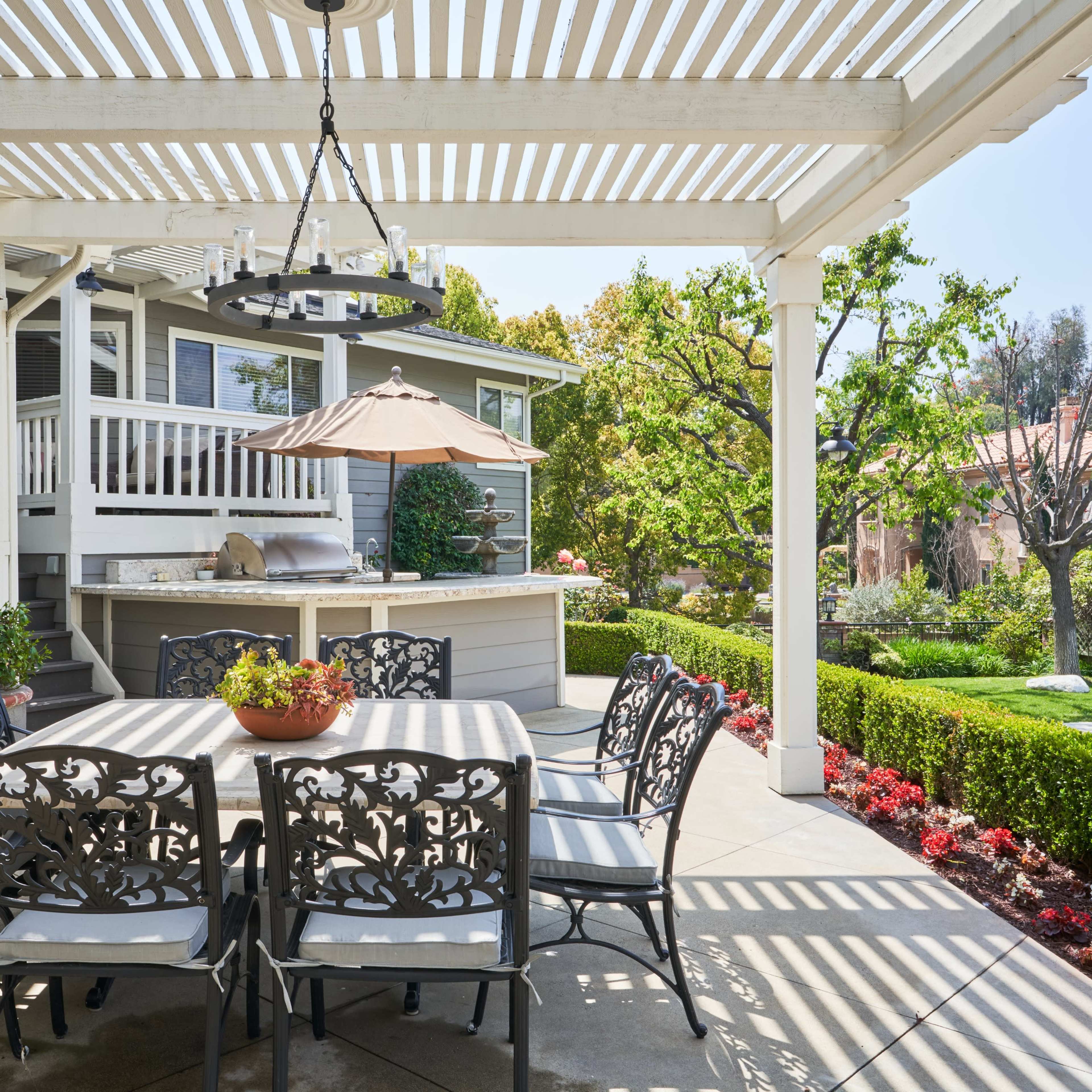 A patio area features a table with six chairs under a pergola, surrounded by well-maintained gardens and a bar space nearby.