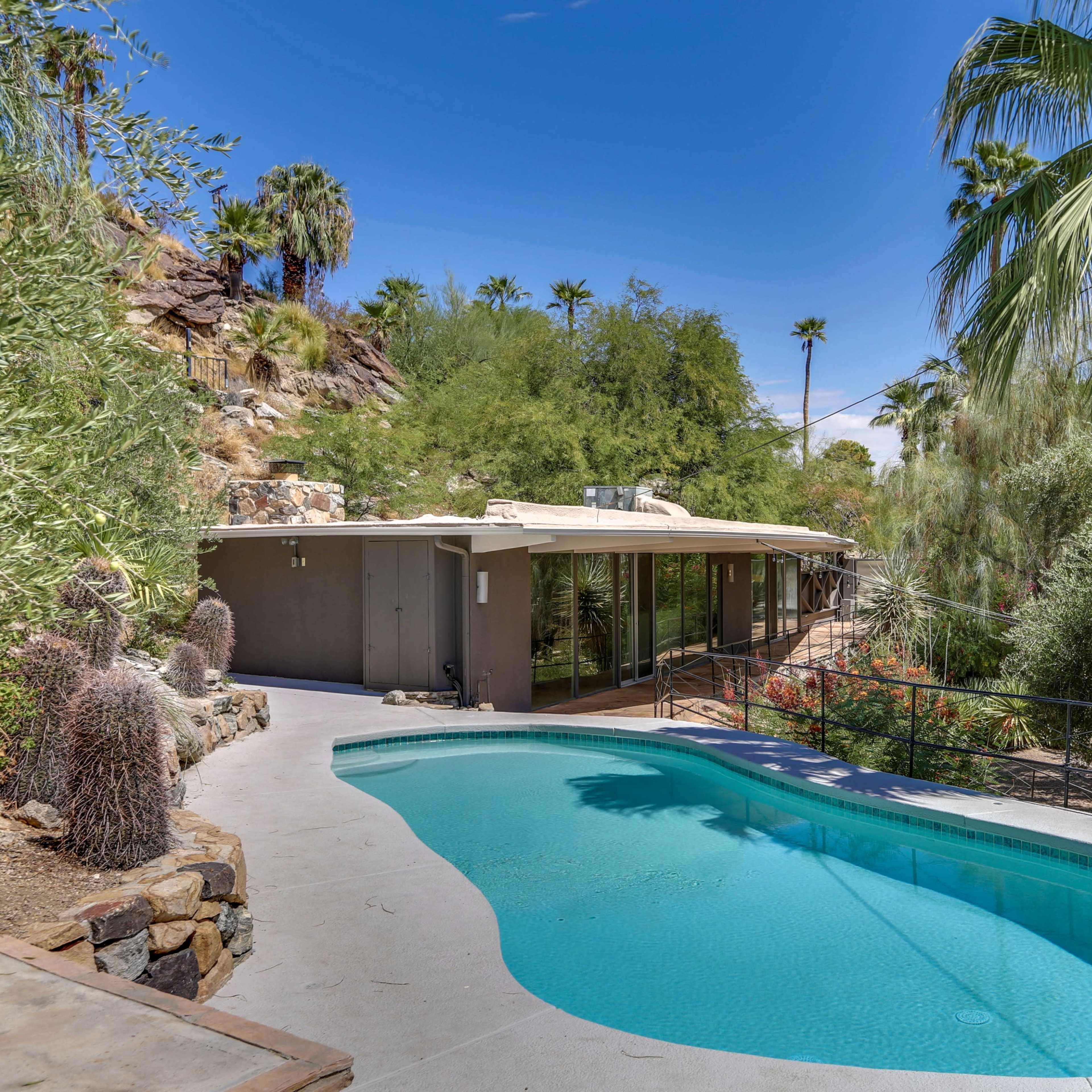 A modern home with large windows overlooks a swimming pool surrounded by palm trees and desert landscaping.
