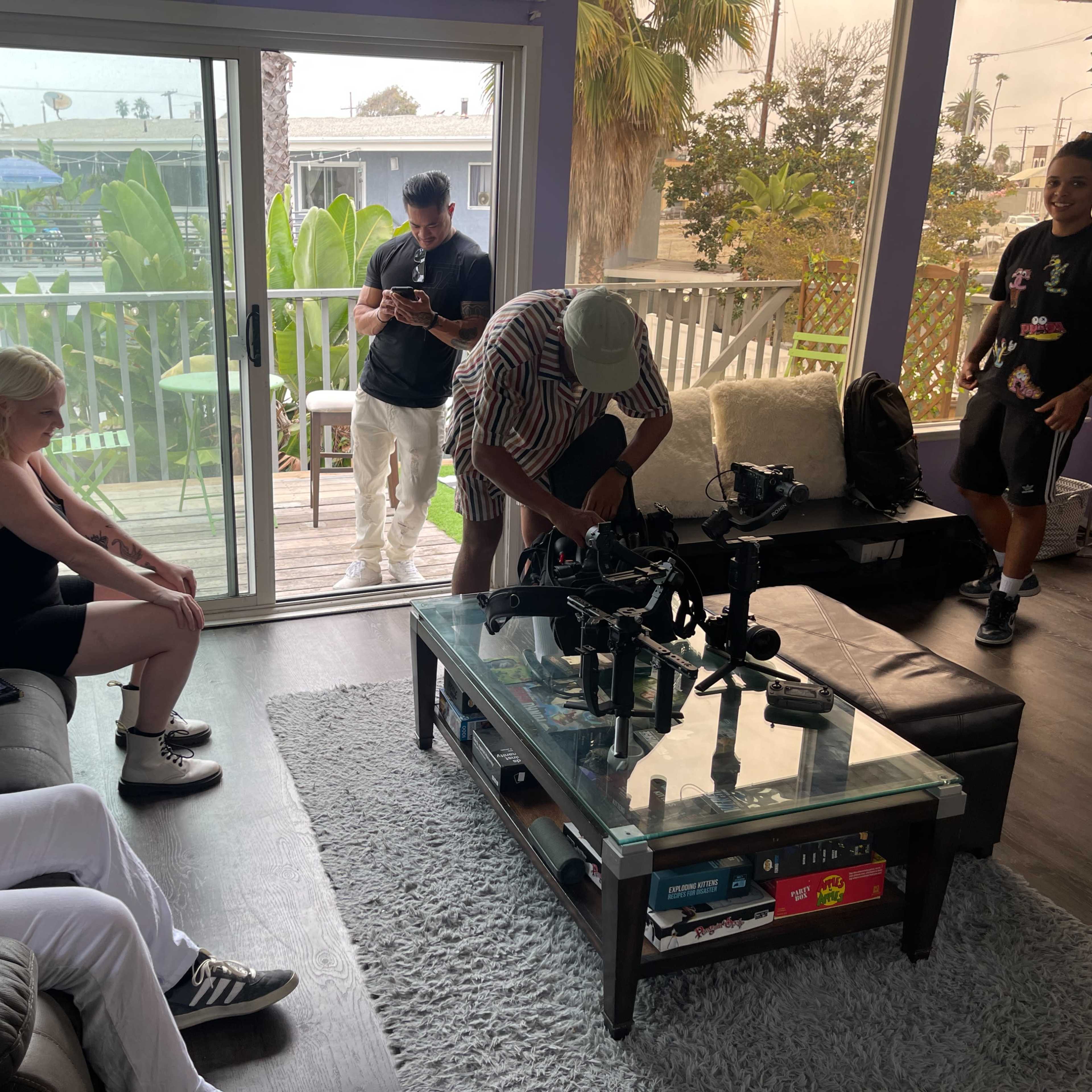 A group of people are gathered in a living room with a glass coffee table, where one person is adjusting camera equipment while others are on their phones.