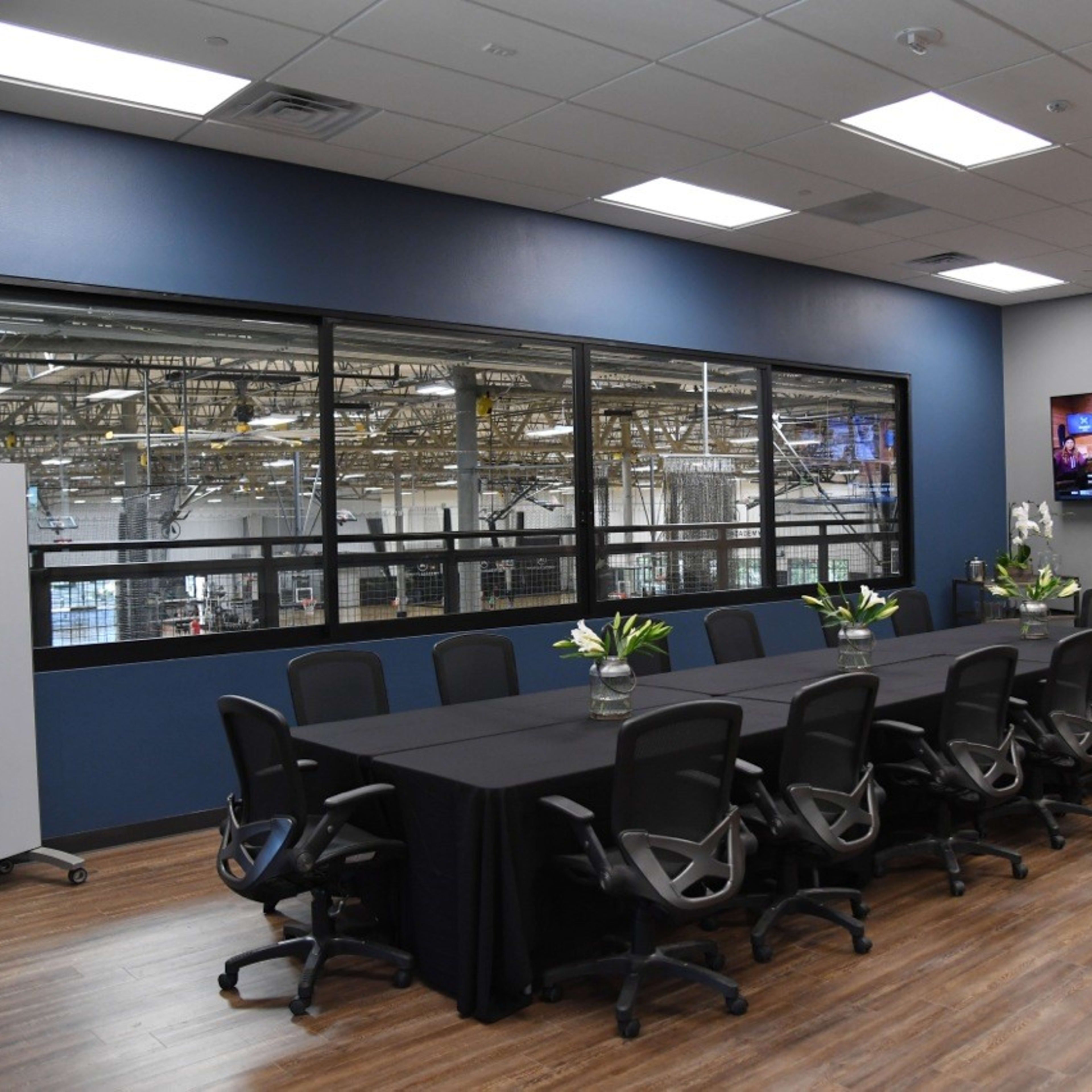 A large conference room features a long table surrounded by office chairs, with a view of a warehouse through large windows and a television mounted on the wall.
