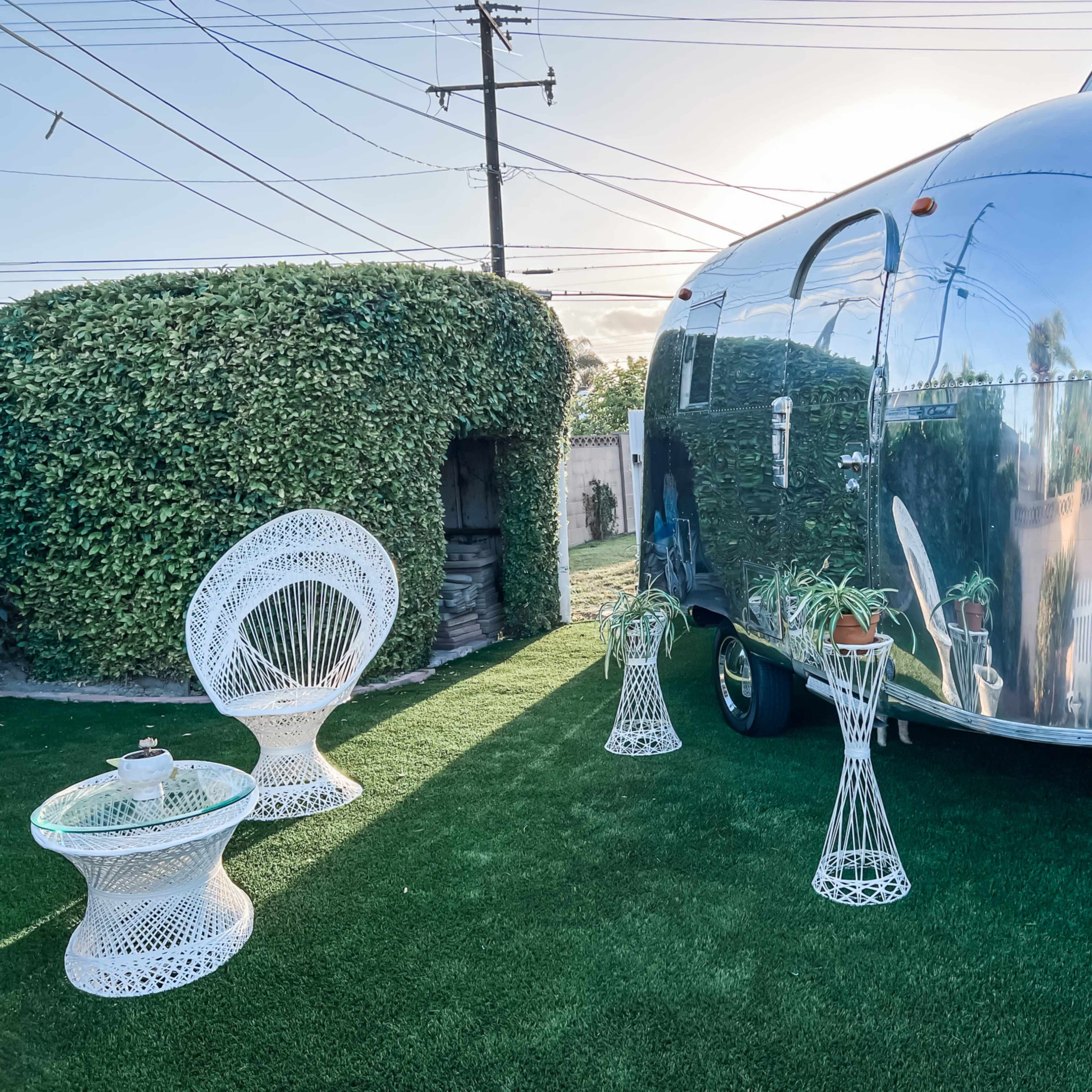 An Airstream trailer sits on green artificial grass beside two white wicker chairs and a decorative table, with a hedged structure in the background.