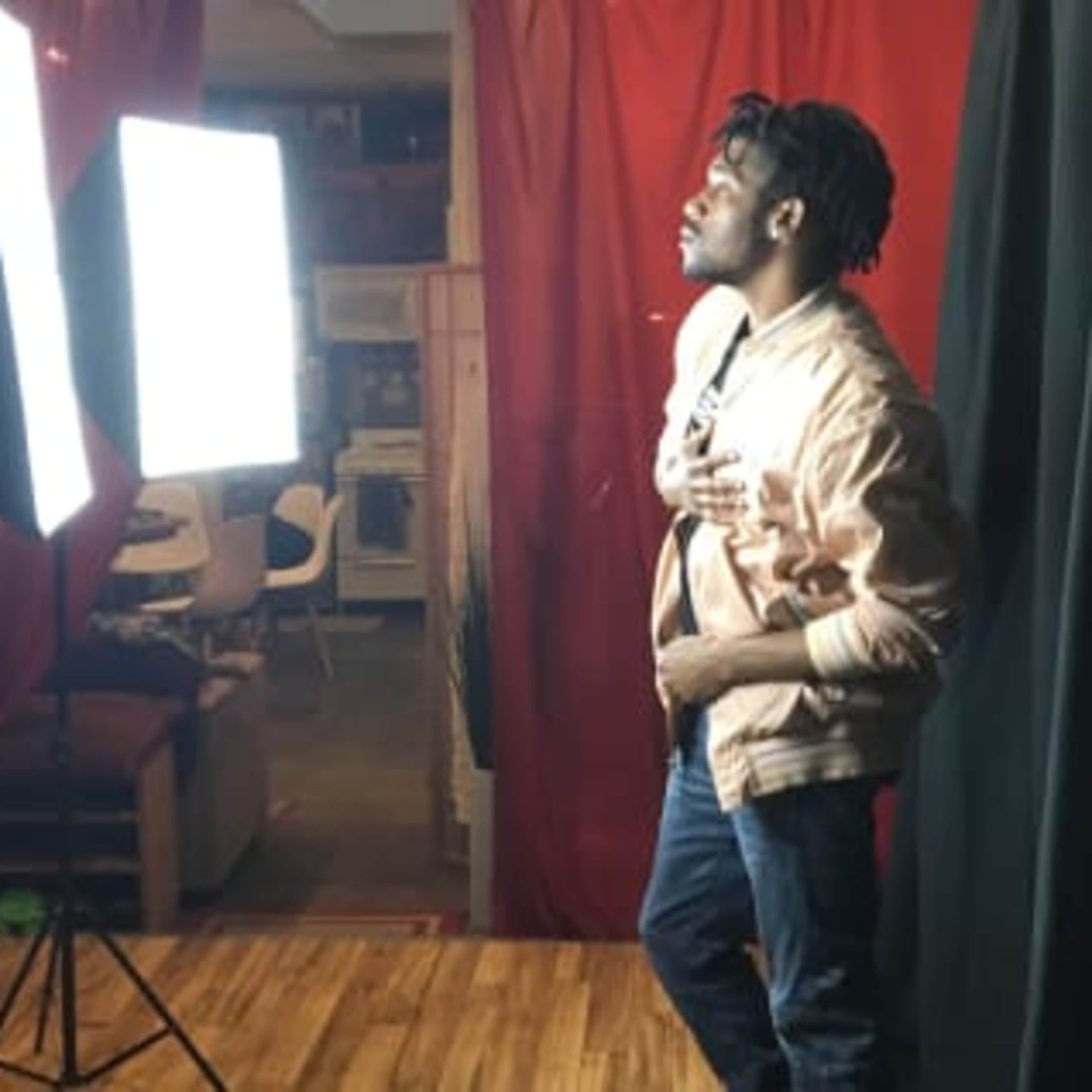A man stands near two bright studio lights, posing in front of a red backdrop.