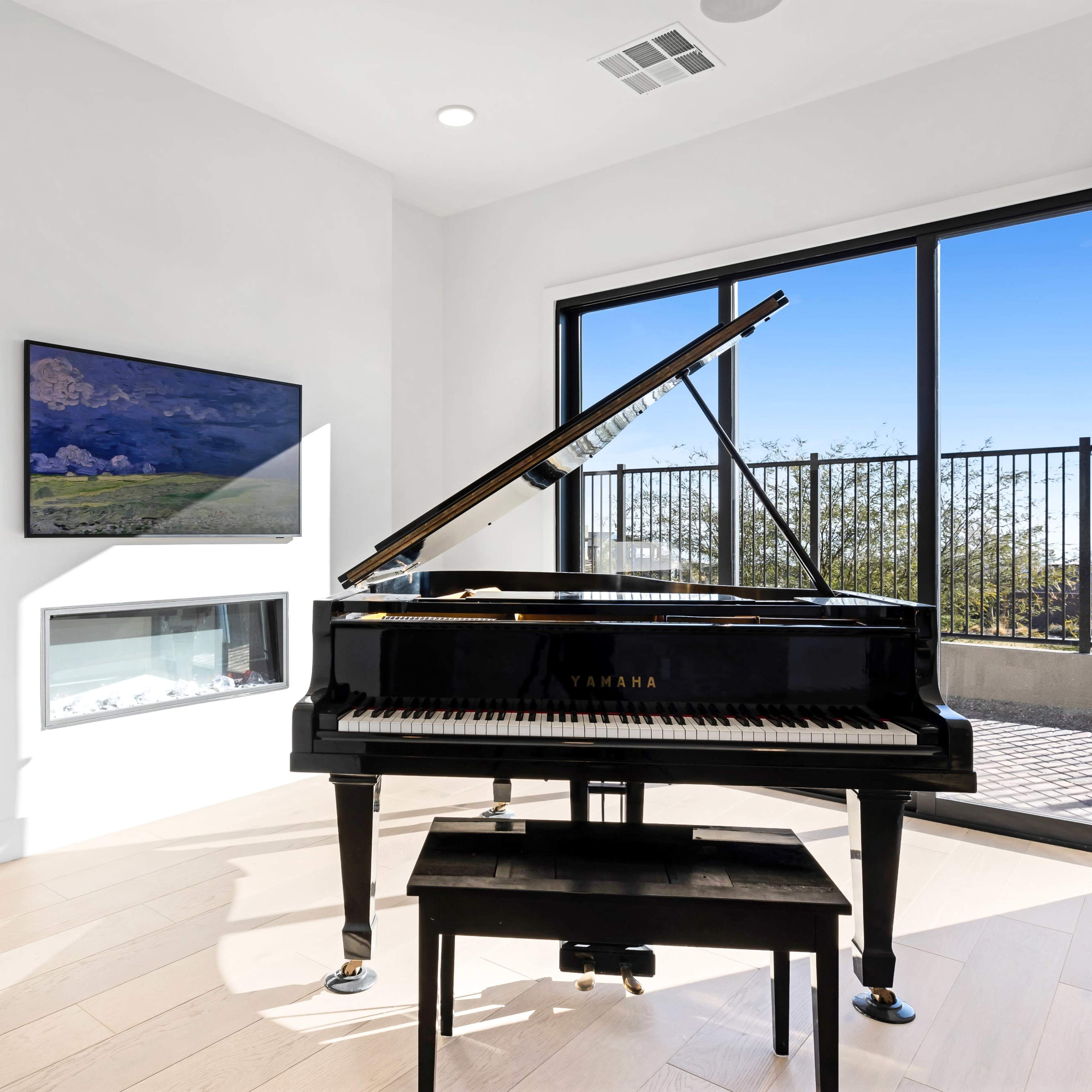 A black Yamaha grand piano is positioned in front of a large window with a view, next to a mounted television and a fireplace.