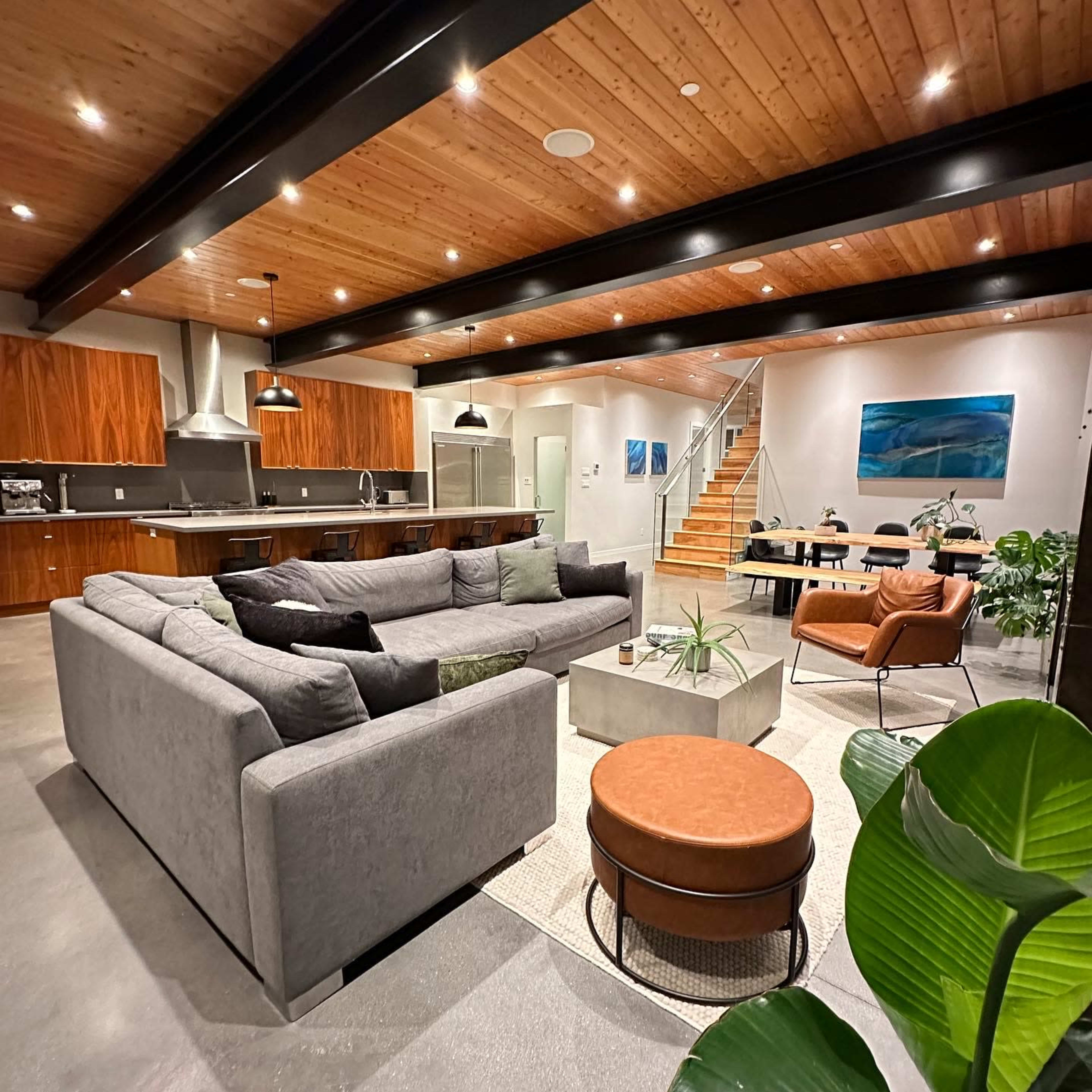 A modern open-concept living space with a large grey sectional sofa, a circular orange ottoman, a dining area, and a kitchen with wooden cabinetry and stainless steel appliances.