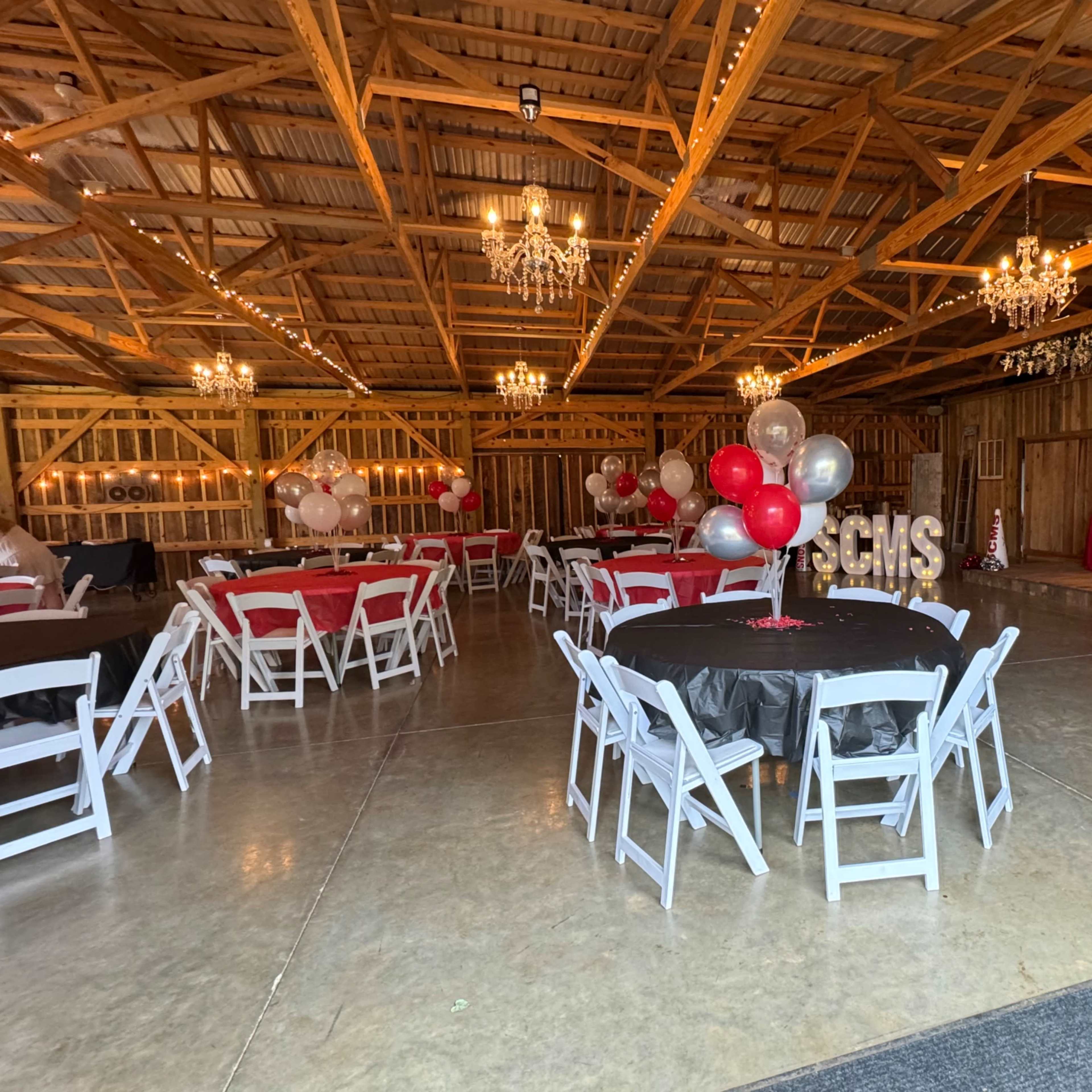The interior of a barn is set up for an event with round tables, balloons, and chandeliers.