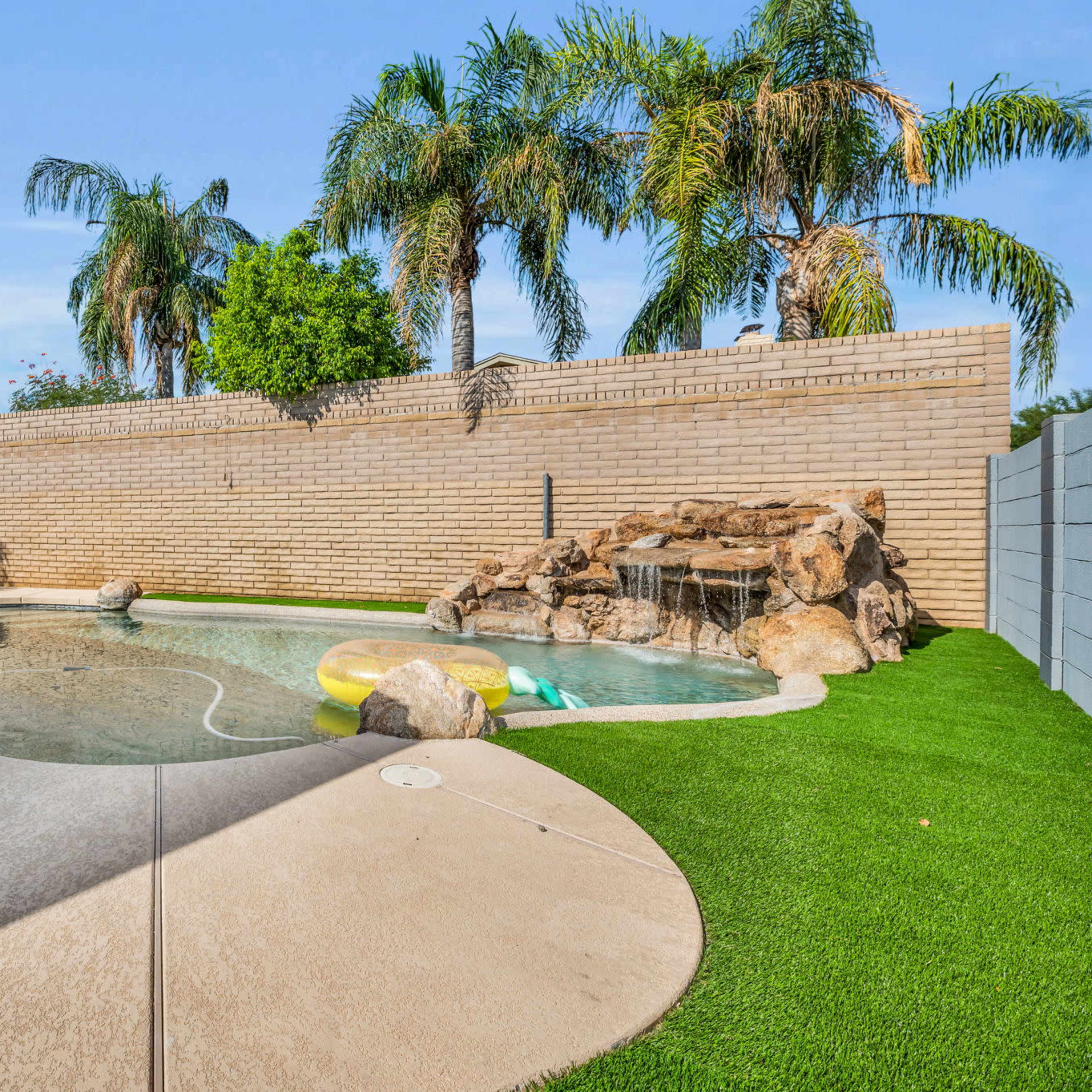 A backyard with a swimming pool, palm trees, a rock waterfall, and a grassy area.