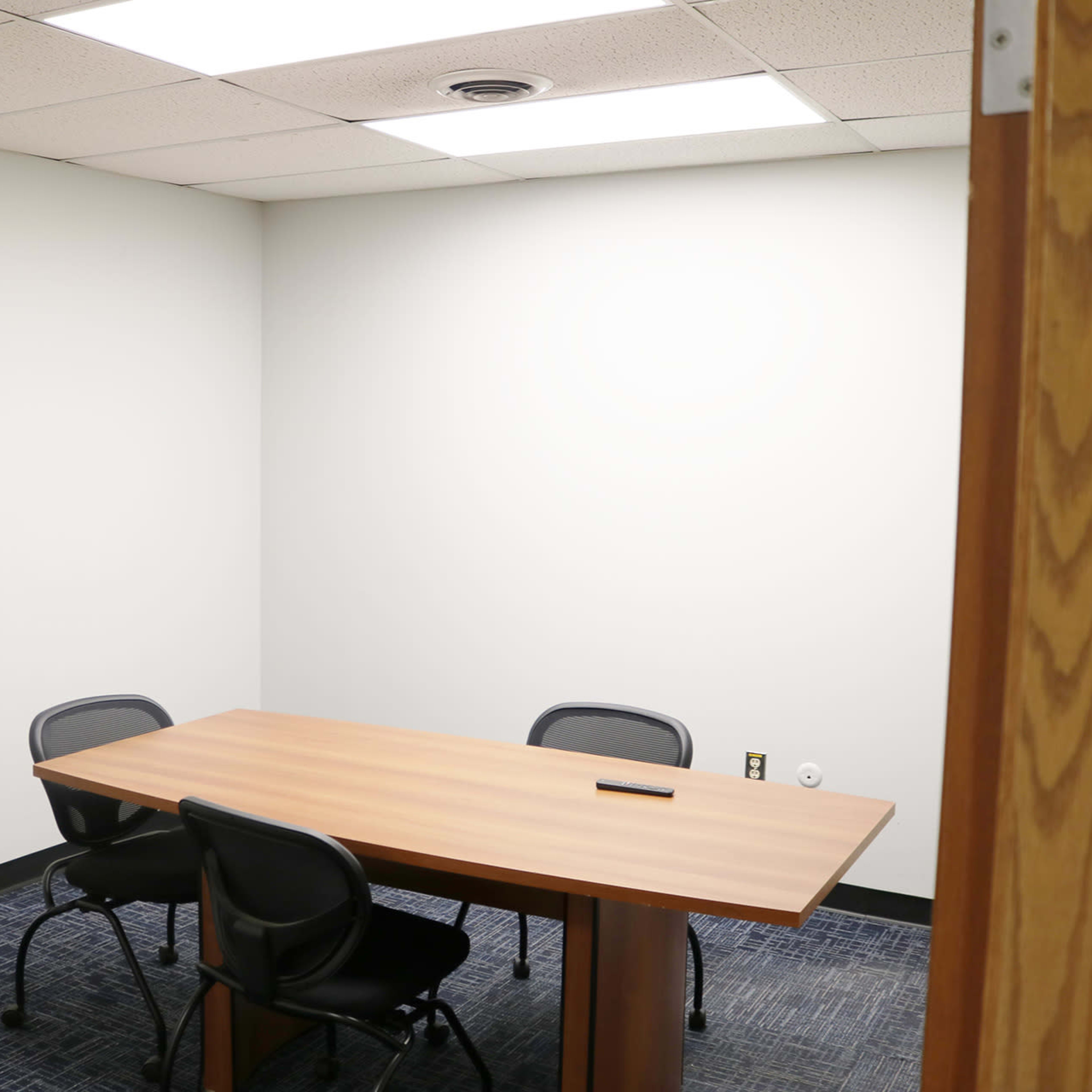 A small, empty meeting room features a wooden table and four chairs against plain walls.