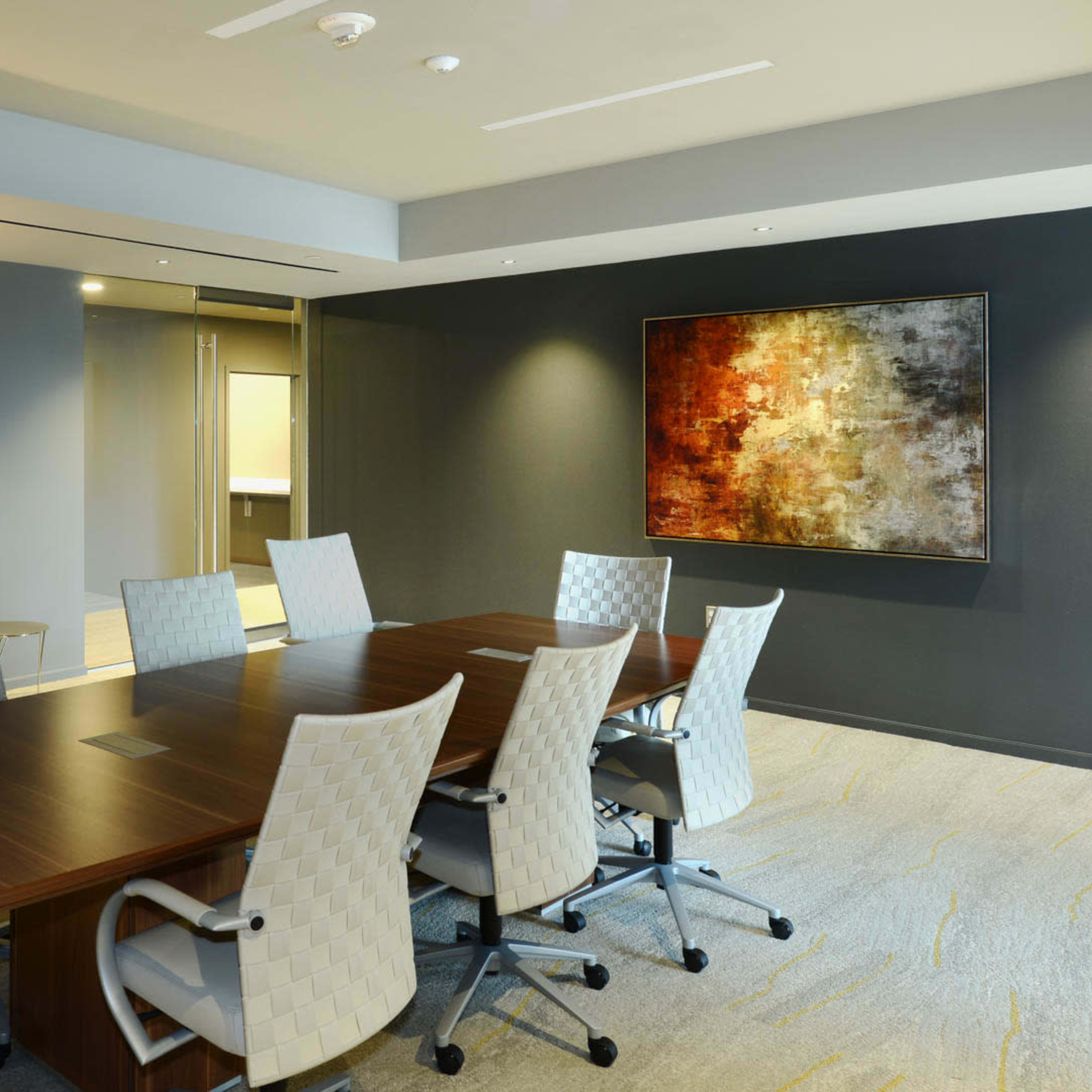 A modern conference room features a large wooden table surrounded by eight ergonomic chairs, with abstract artwork on the walls and a lamp in one corner.