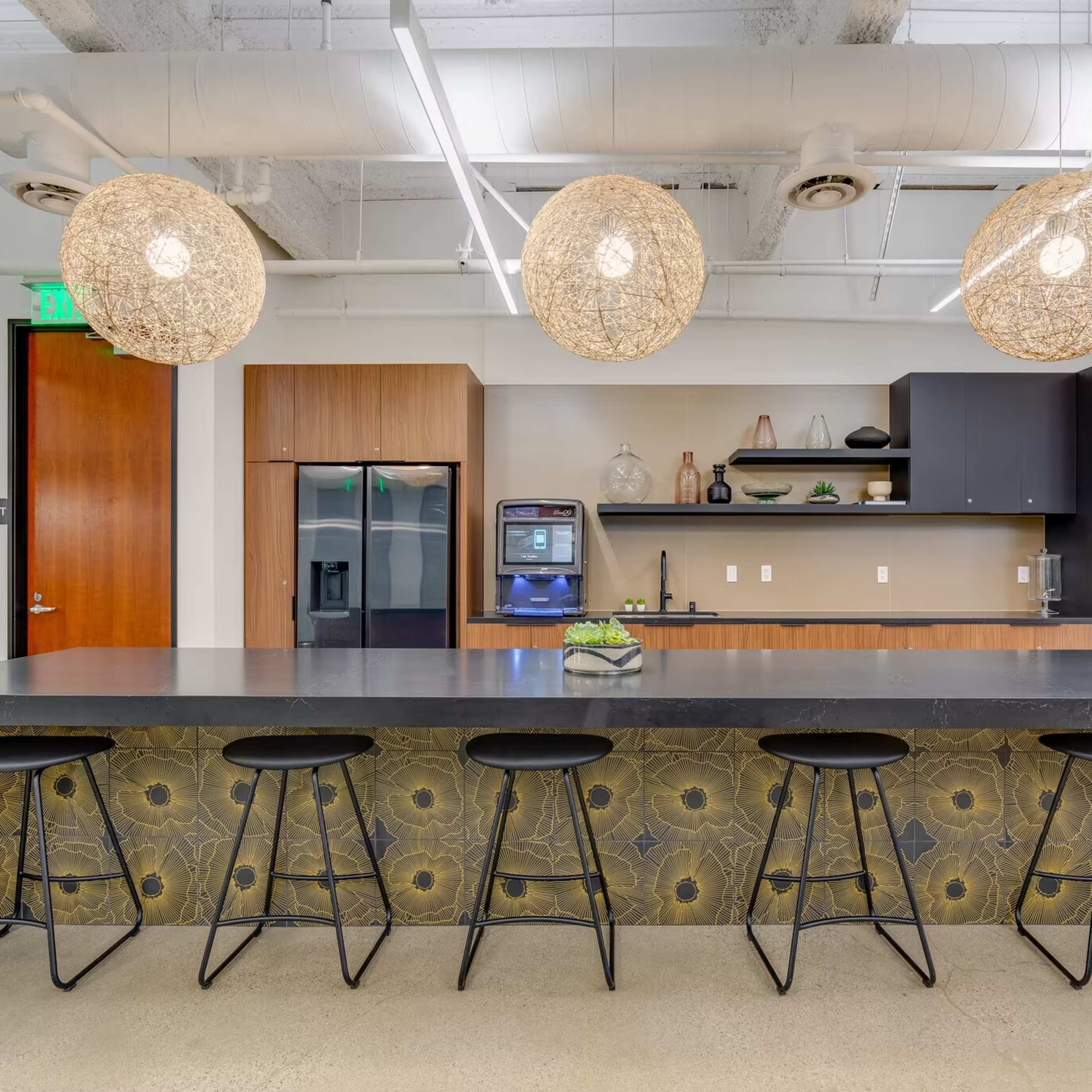 A modern kitchen with a large dark countertop, bar stools, and stylish pendant lights overhead.
