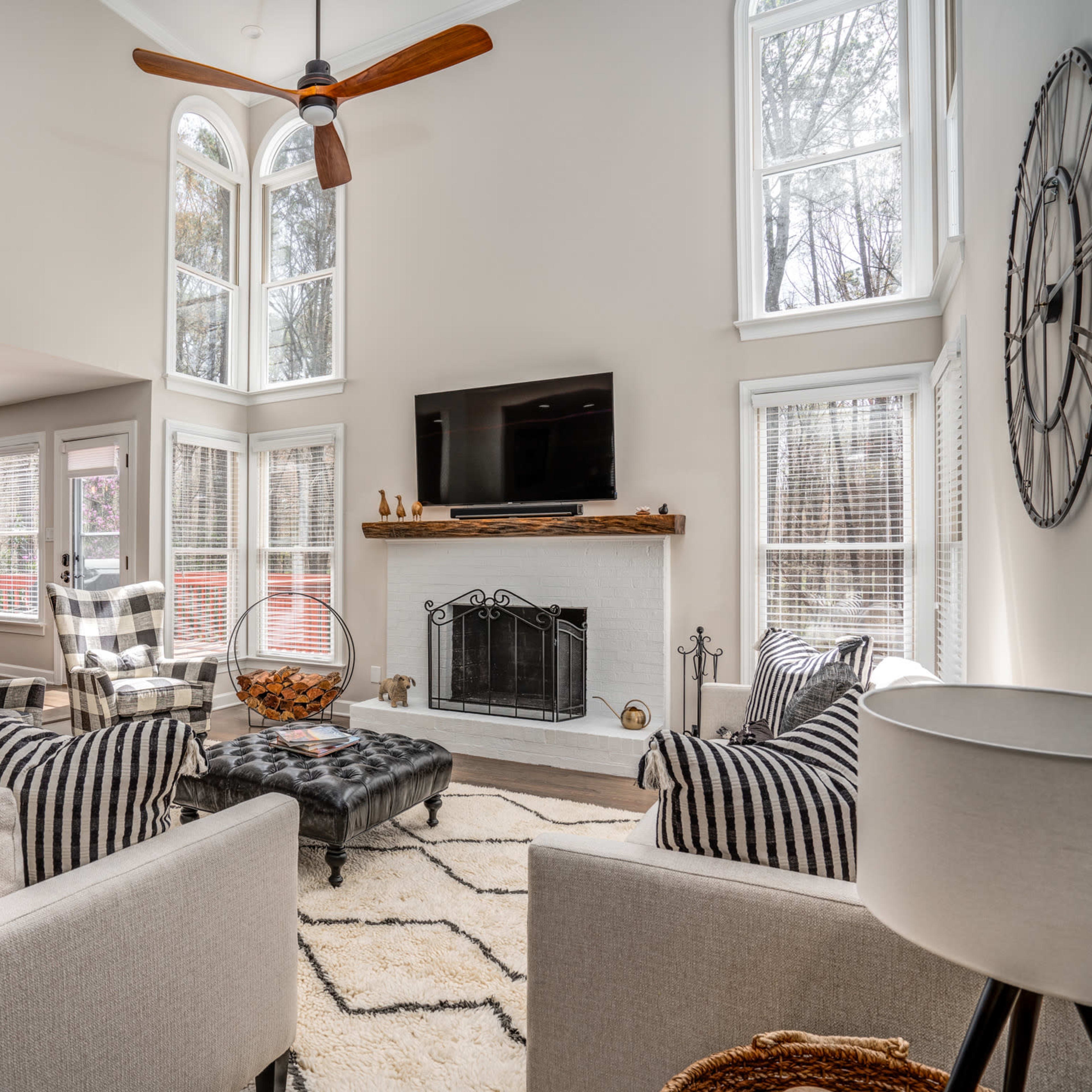 A modern living room features a high ceiling, large windows, a central fireplace, and a mix of patterned furniture arranged around a coffee table.