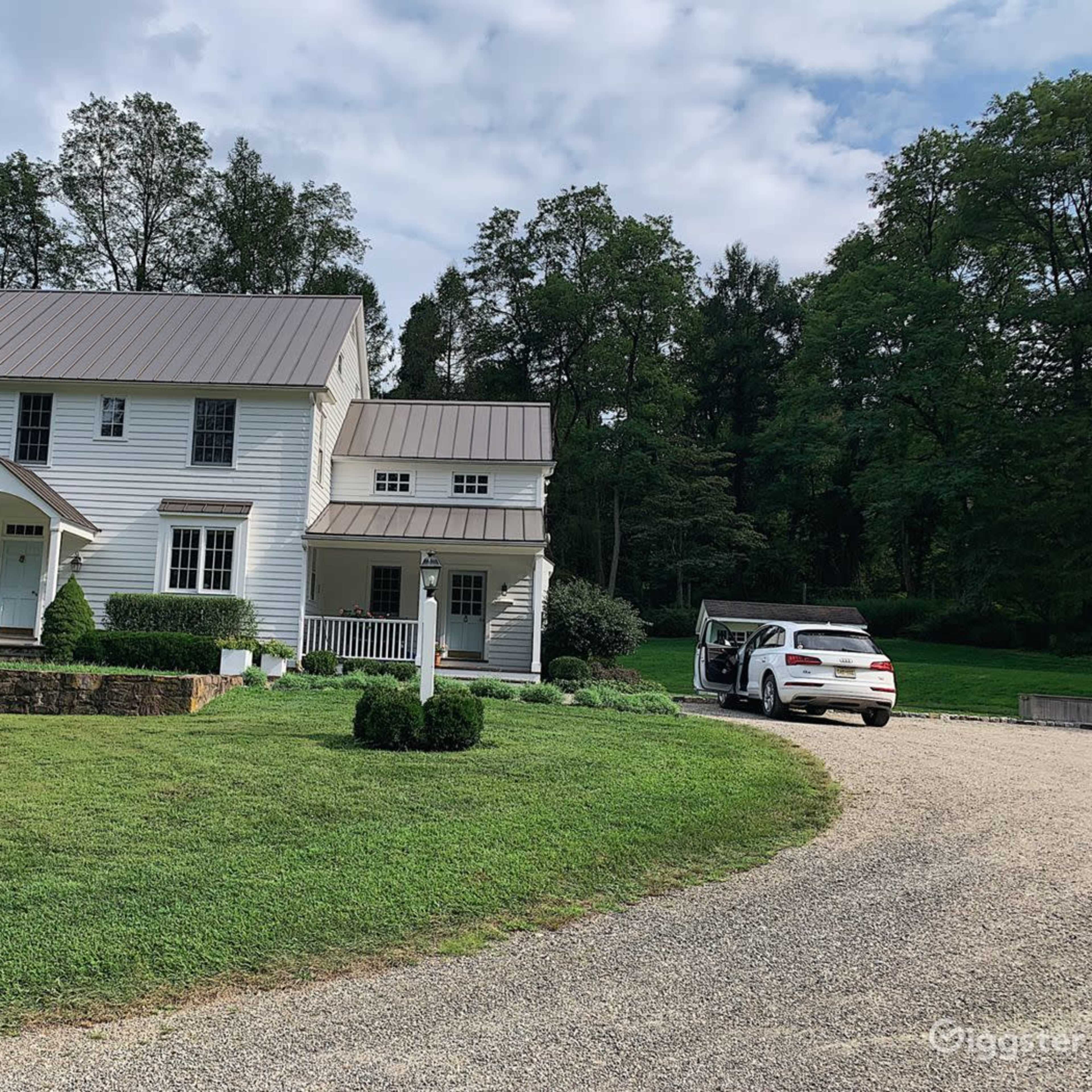 A two-story white house with a metal roof is surrounded by grassy areas and trees, with a gravel driveway leading to a parked car.
