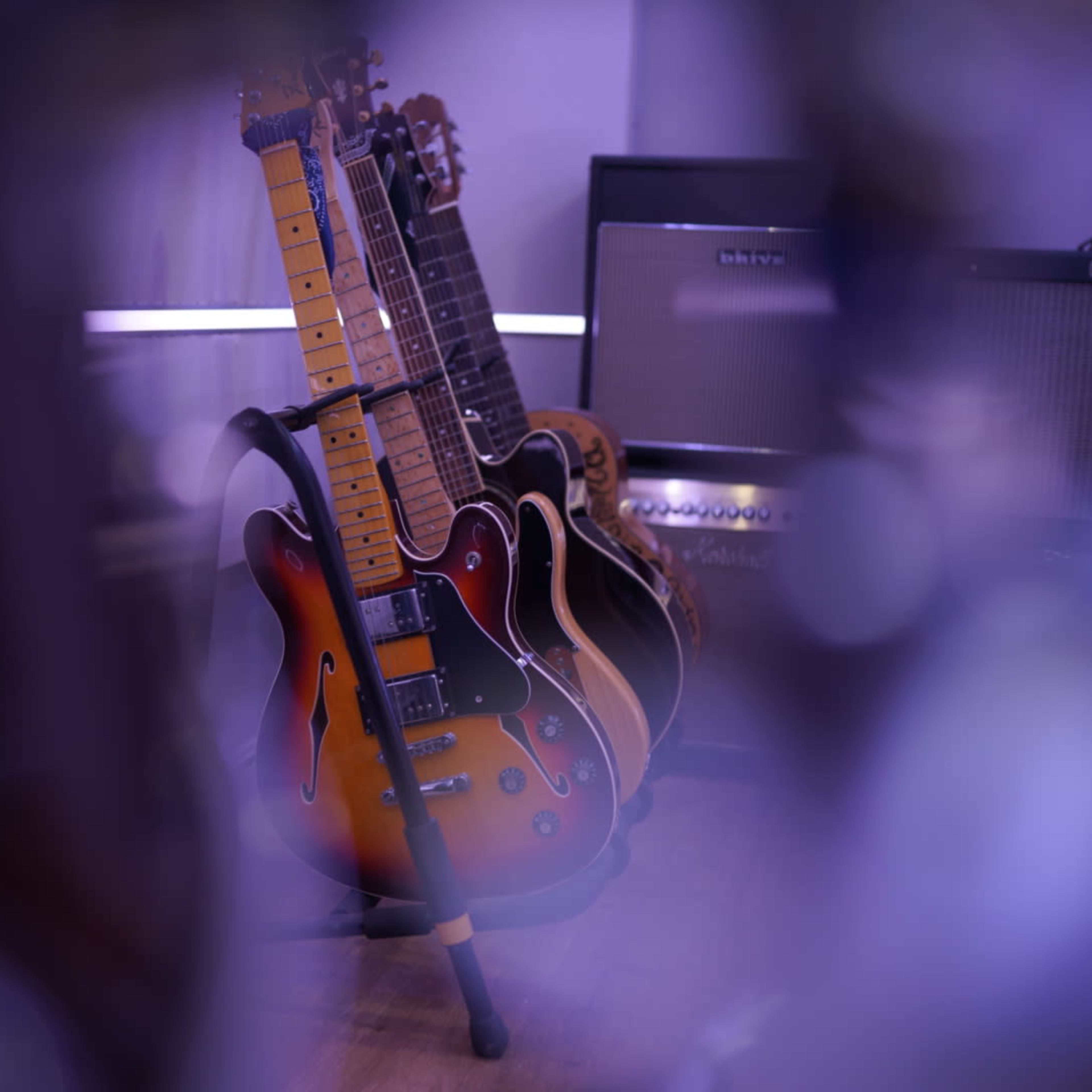 A collection of guitars is displayed on stands with amplifiers visible in the background.