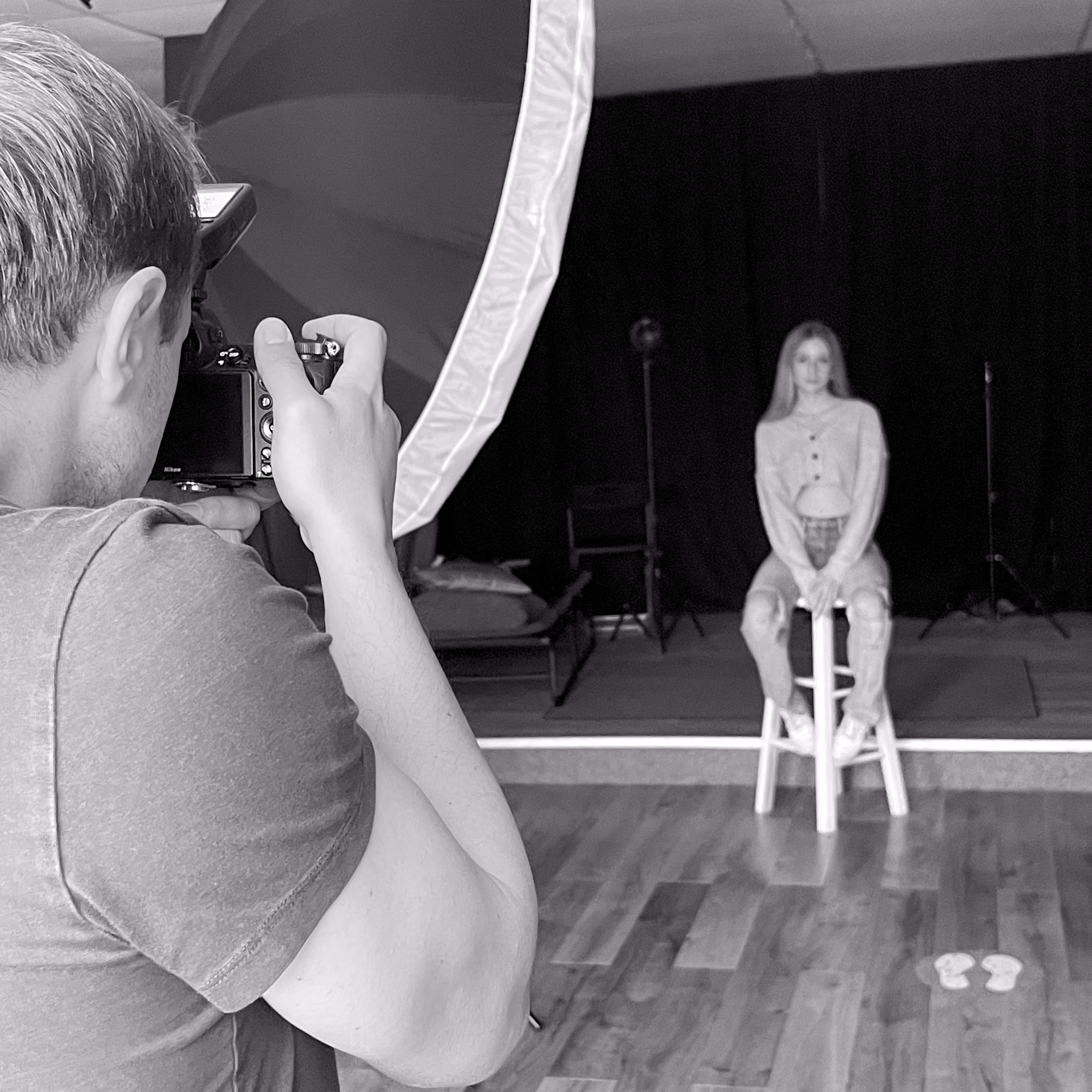 A photographer is taking a picture of a person sitting on a stool in a studio with a large umbrella softbox in the foreground.