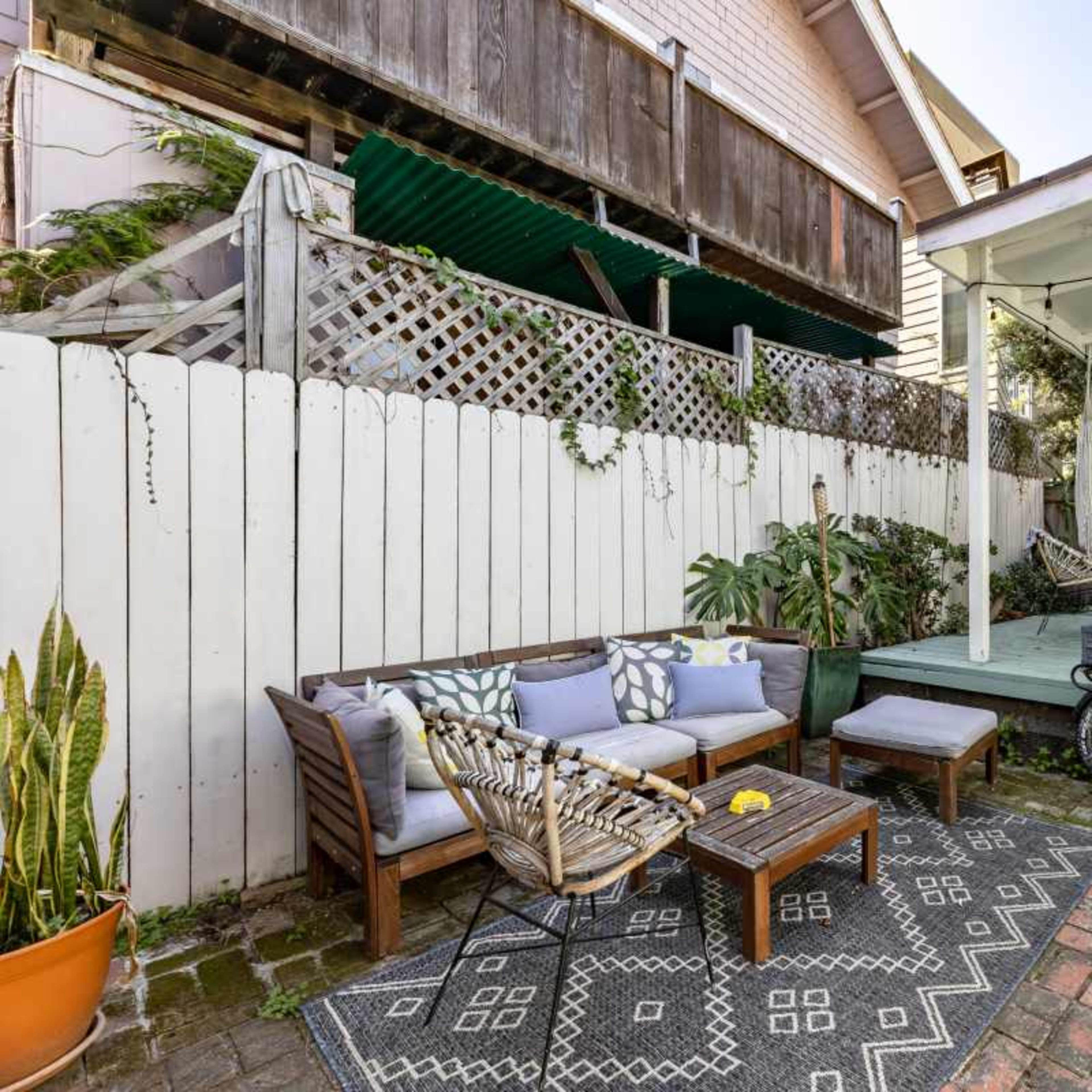 A small backyard featuring a seating area with a wooden couch and a coffee table, surrounded by potted plants and a bicycle parked near a covered patio.