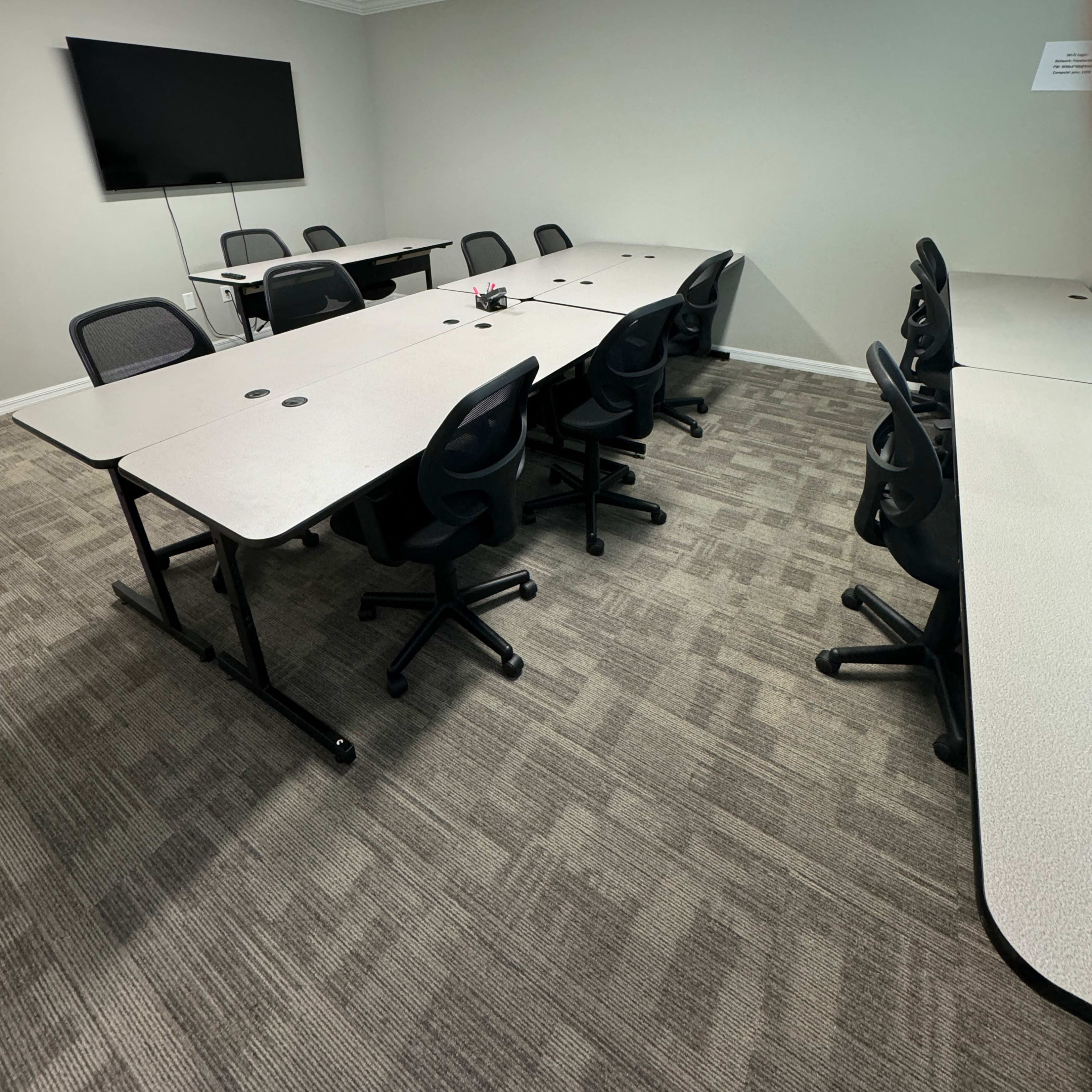 A conference room is shown with several tables arranged in a U-shape, surrounded by black swivel chairs and featuring a wall-mounted TV.
