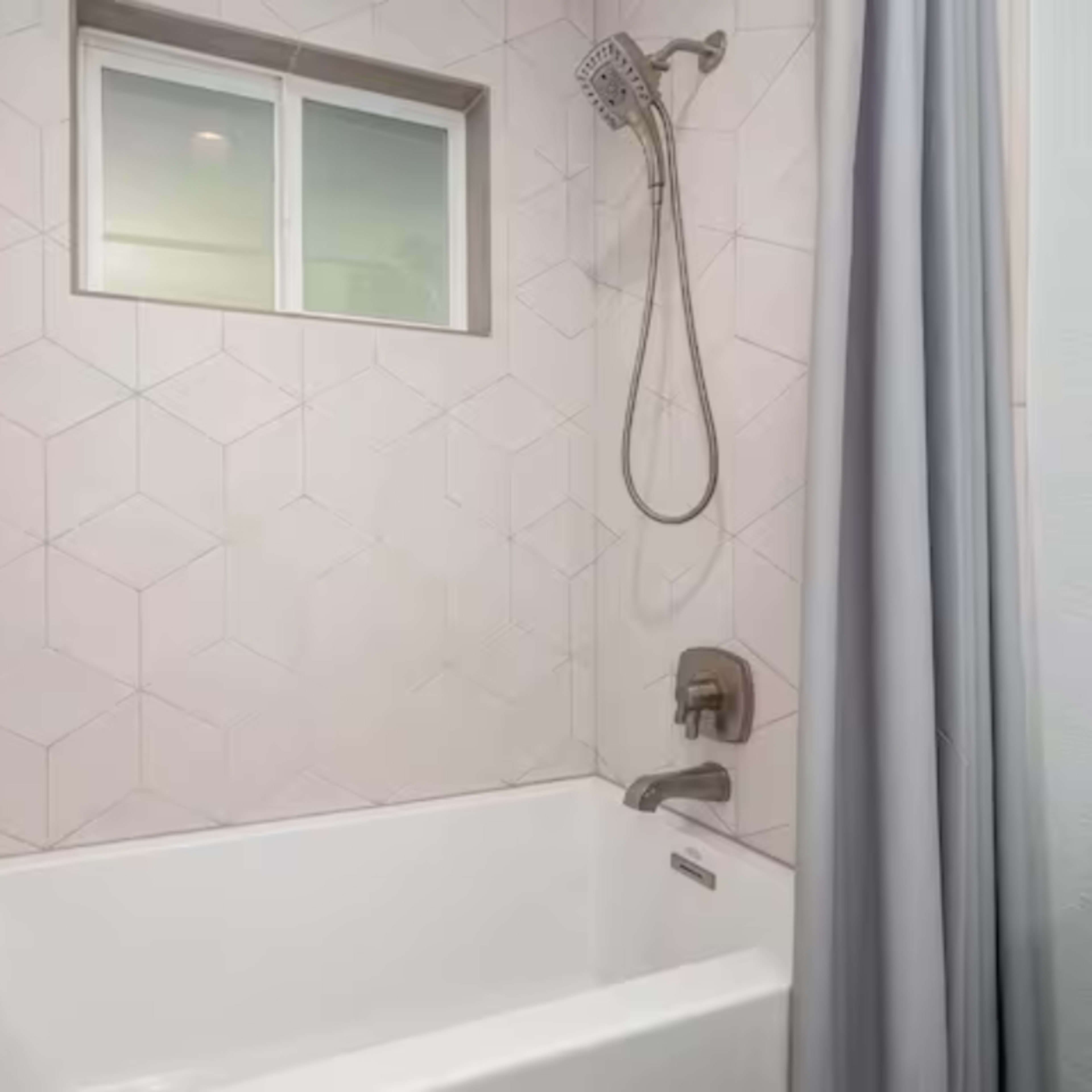 A modern bathroom featuring a white bathtub, a shower with a handheld fixture, and a small window above a geometric tiled wall.