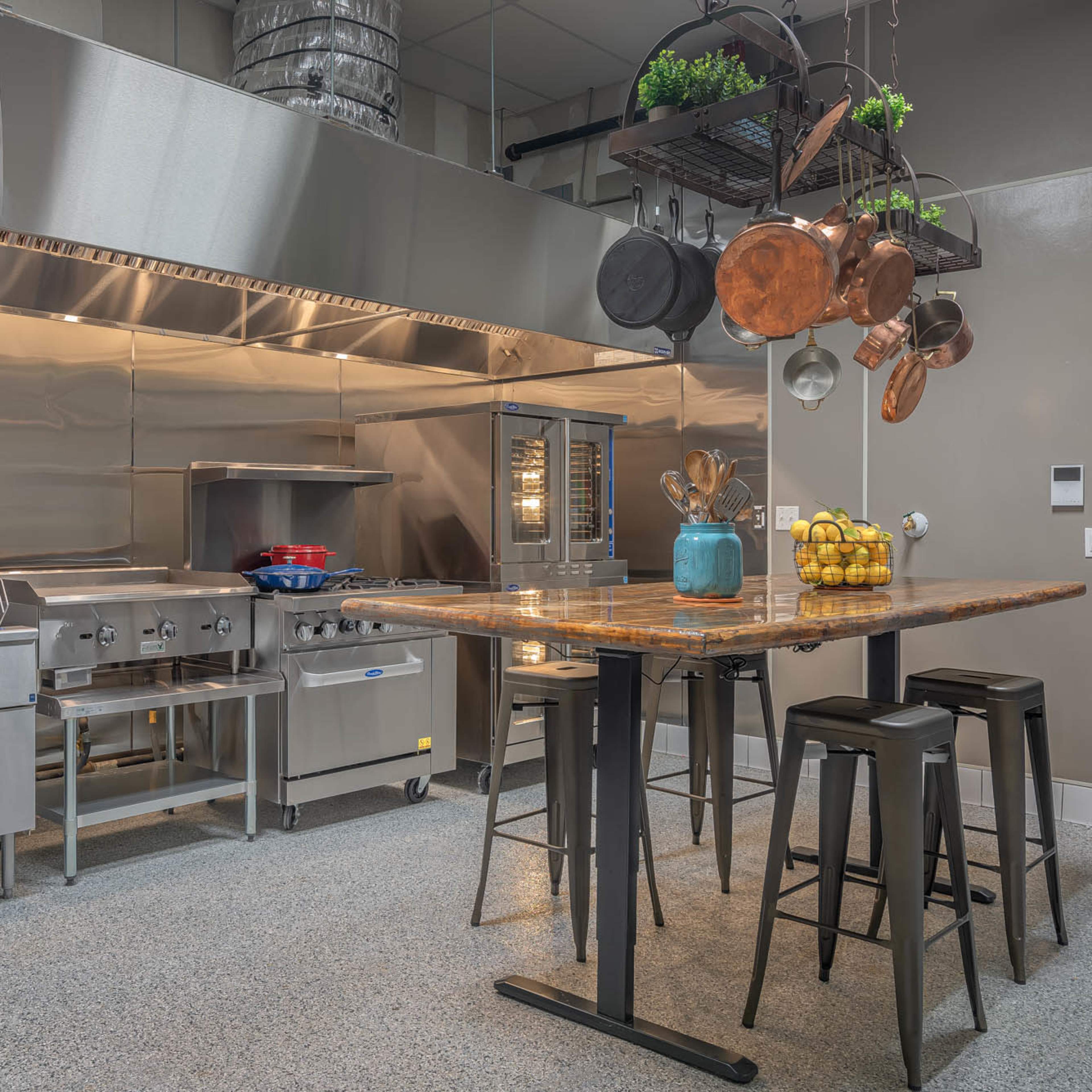 A modern kitchen with stainless steel appliances, a large shared table, and cookware hanging from a rack above.