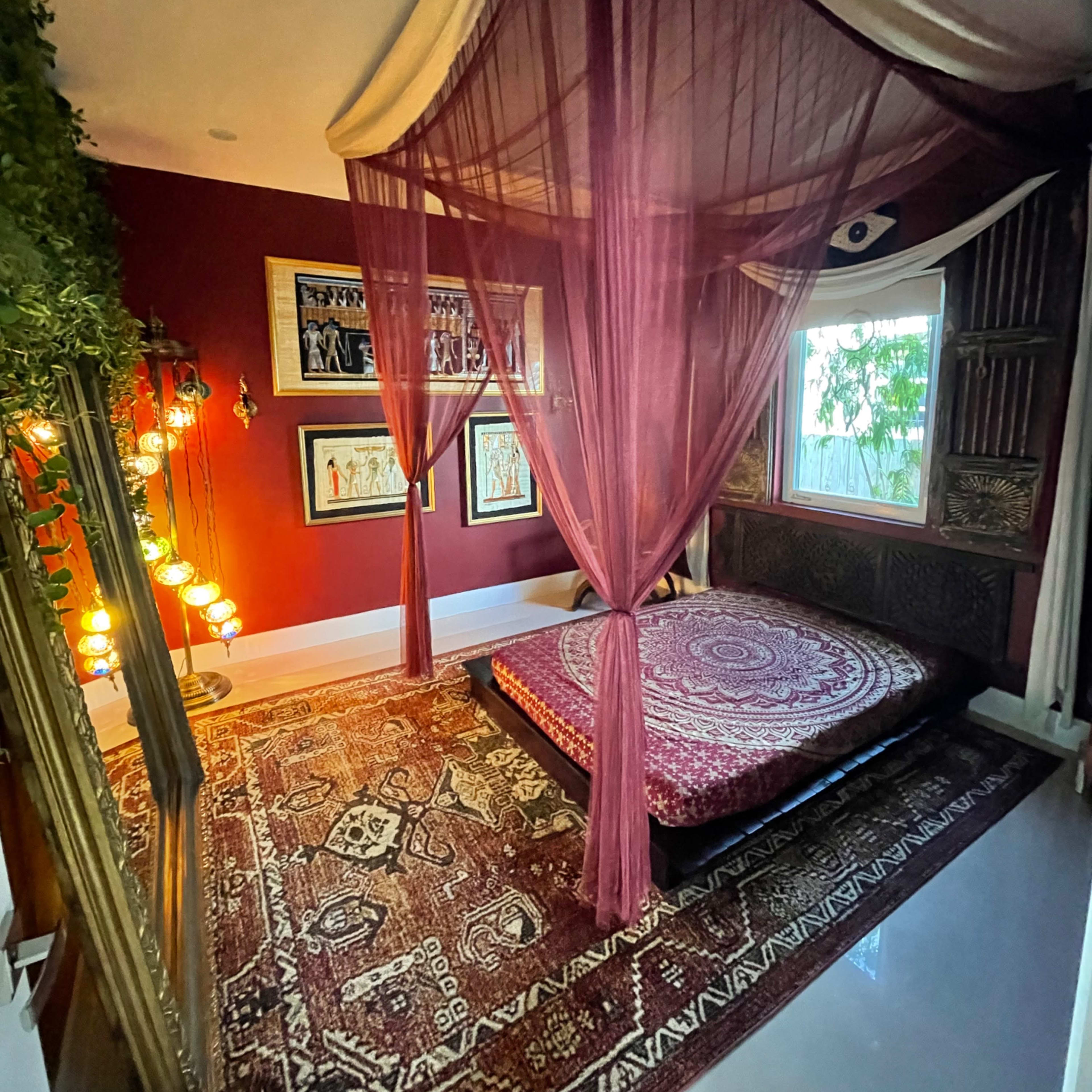 A bedroom features a large, round patterned rug beneath a low, circular mattress draped with sheer curtains, surrounded by decorative wall art and ambient lighting.