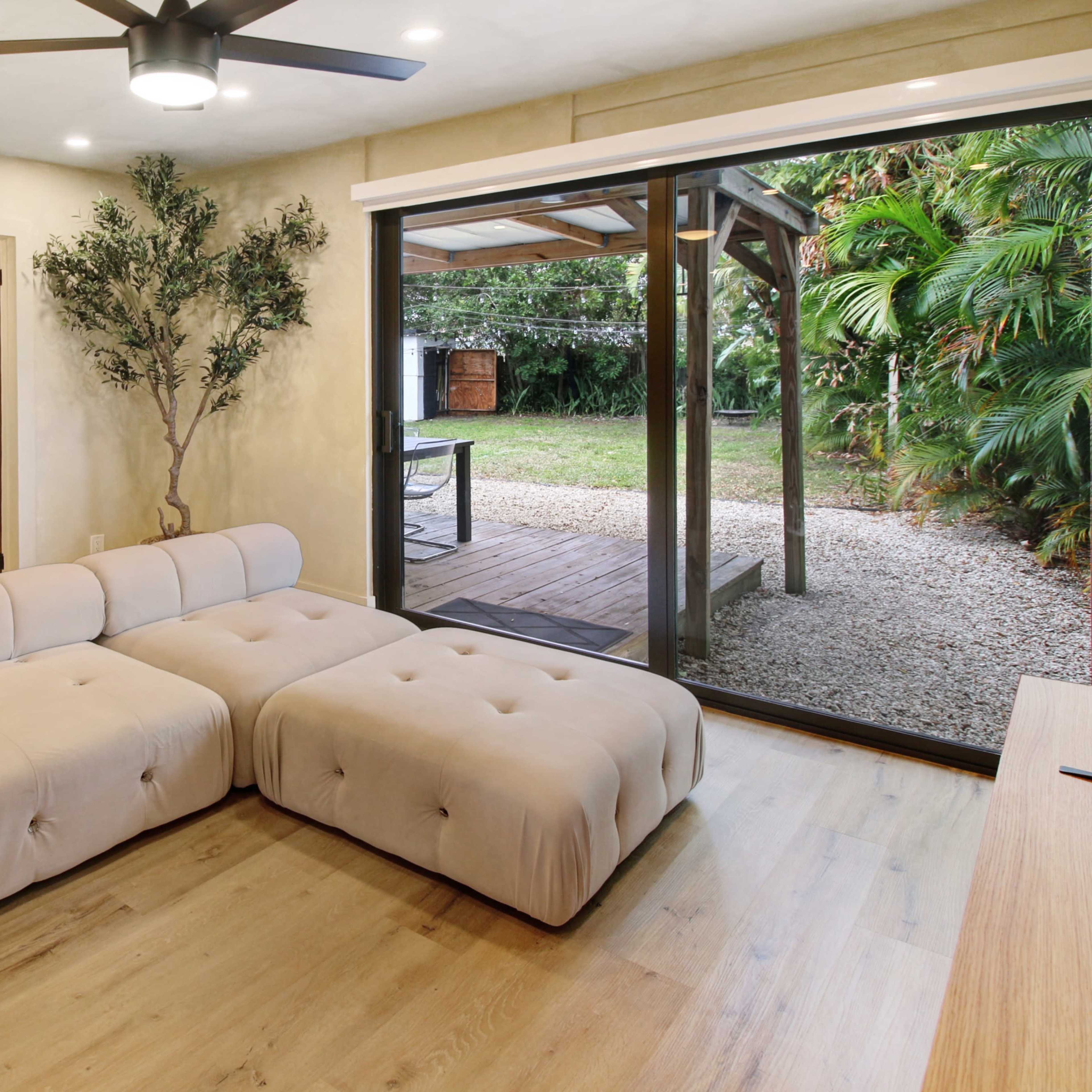 A modern living room features a large beige sectional sofa, a sliding glass door leading to a gravel outdoor area, and a small tree in the corner.