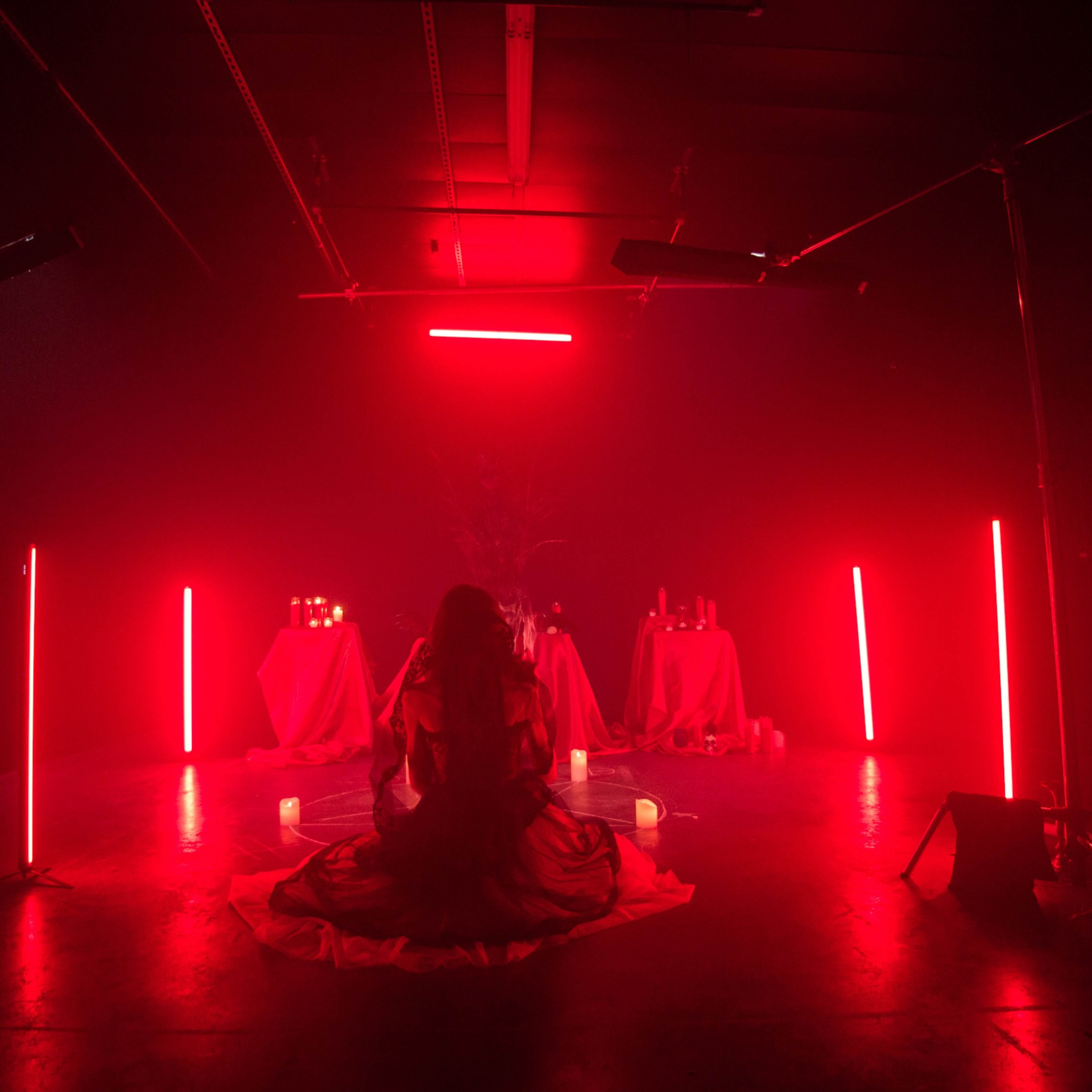A woman sits on a floor surrounded by red-lit tables and candles in a darkened studio.
