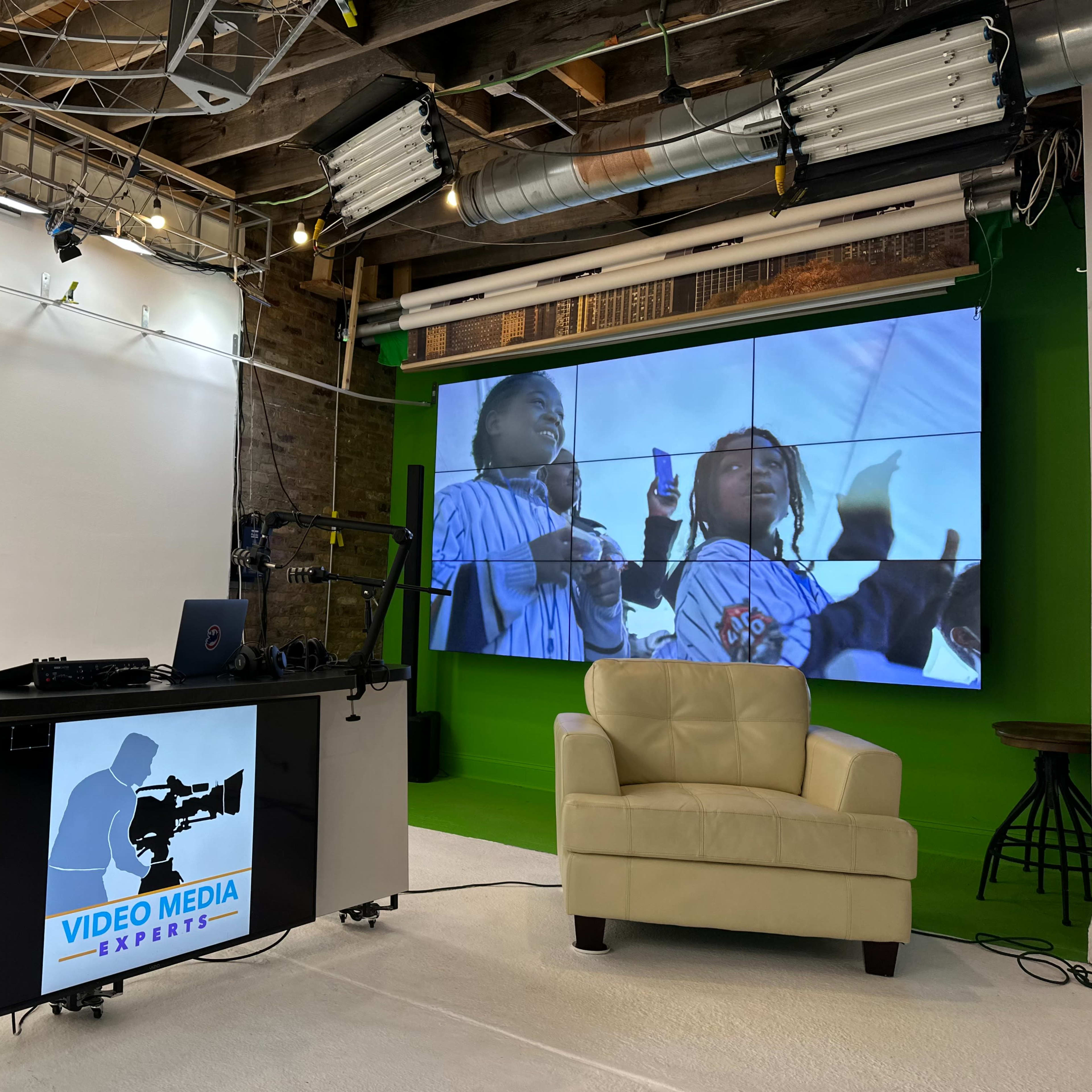 A media studio setup with a large screen displaying two children holding drinks, a cream-colored armchair, and microphones arranged on a black table.