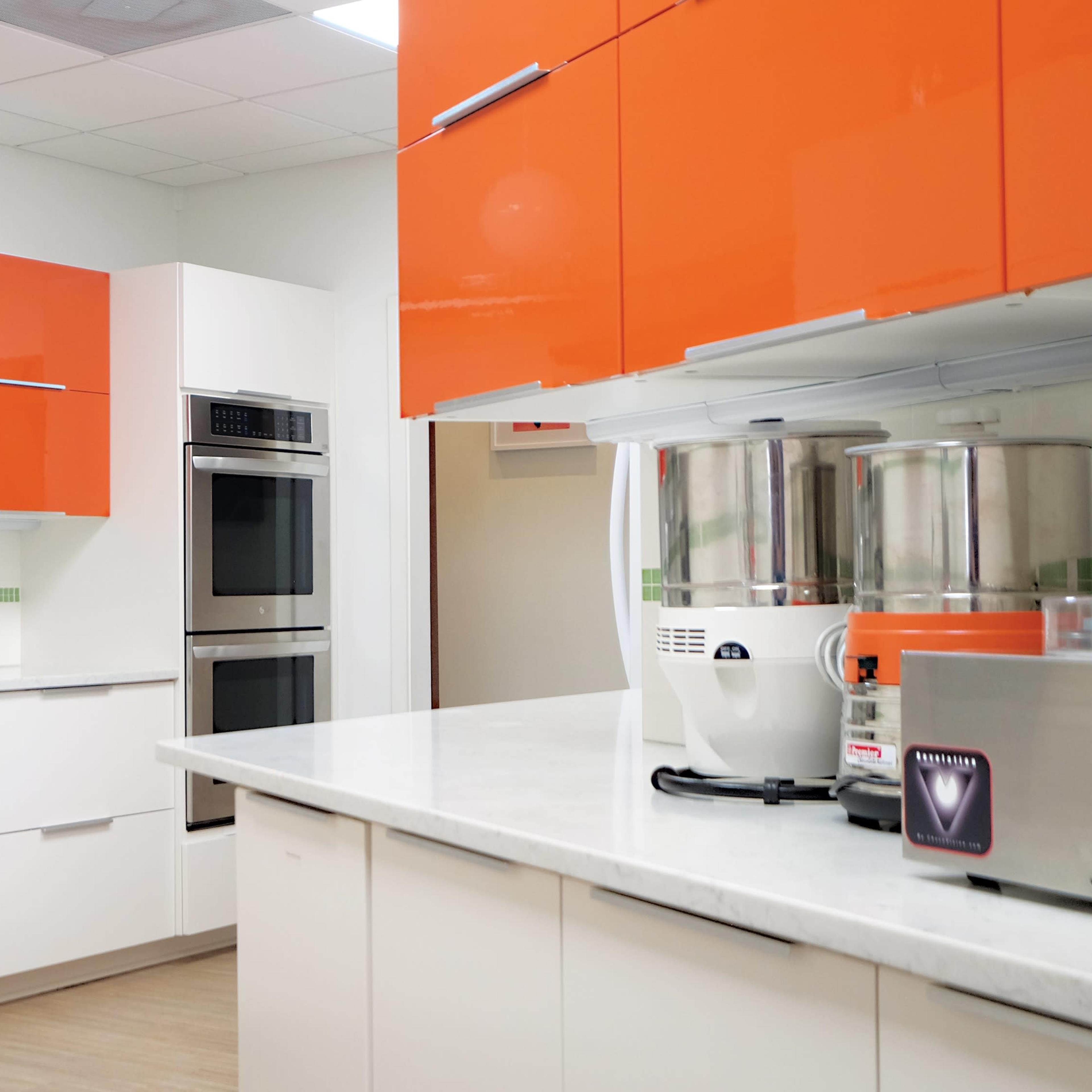 A modern kitchen features orange upper cabinets, stainless steel appliances, and a smooth white countertop.