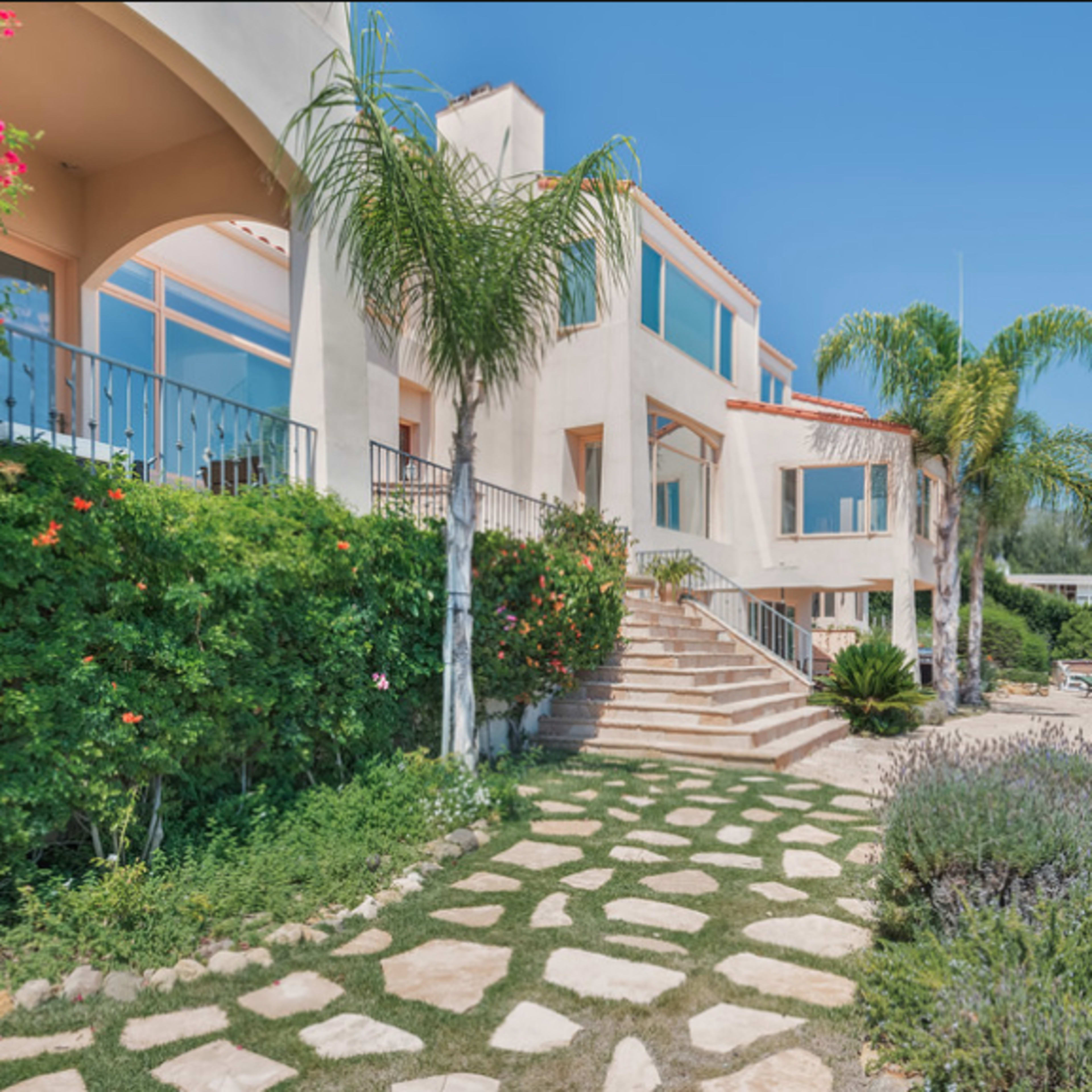 A modern house with a landscaped yard and stone pathway leads to a set of stairs flanked by palm trees.