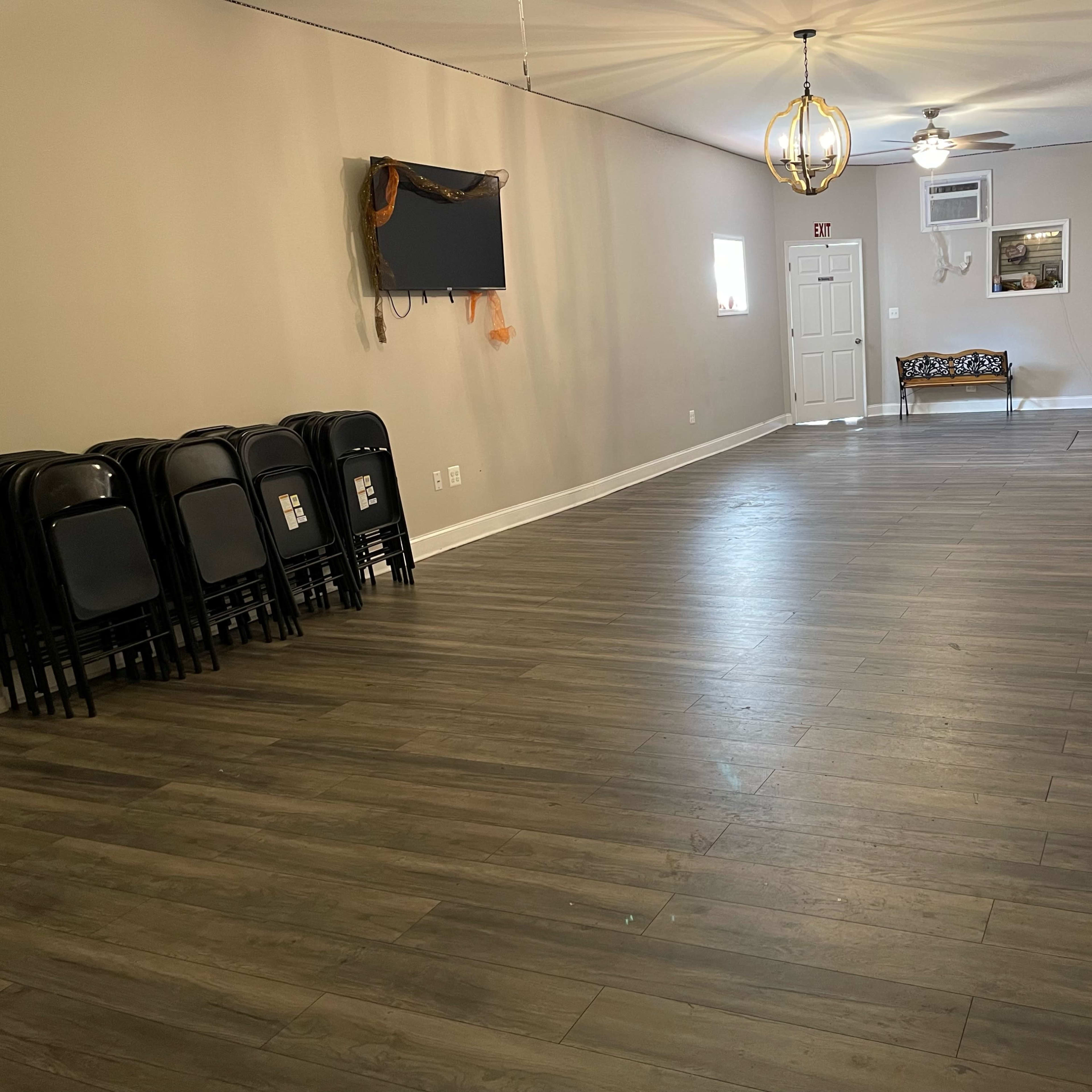 An empty room with gray wooden flooring, a small seating area, and black folding chairs stacked against the wall.