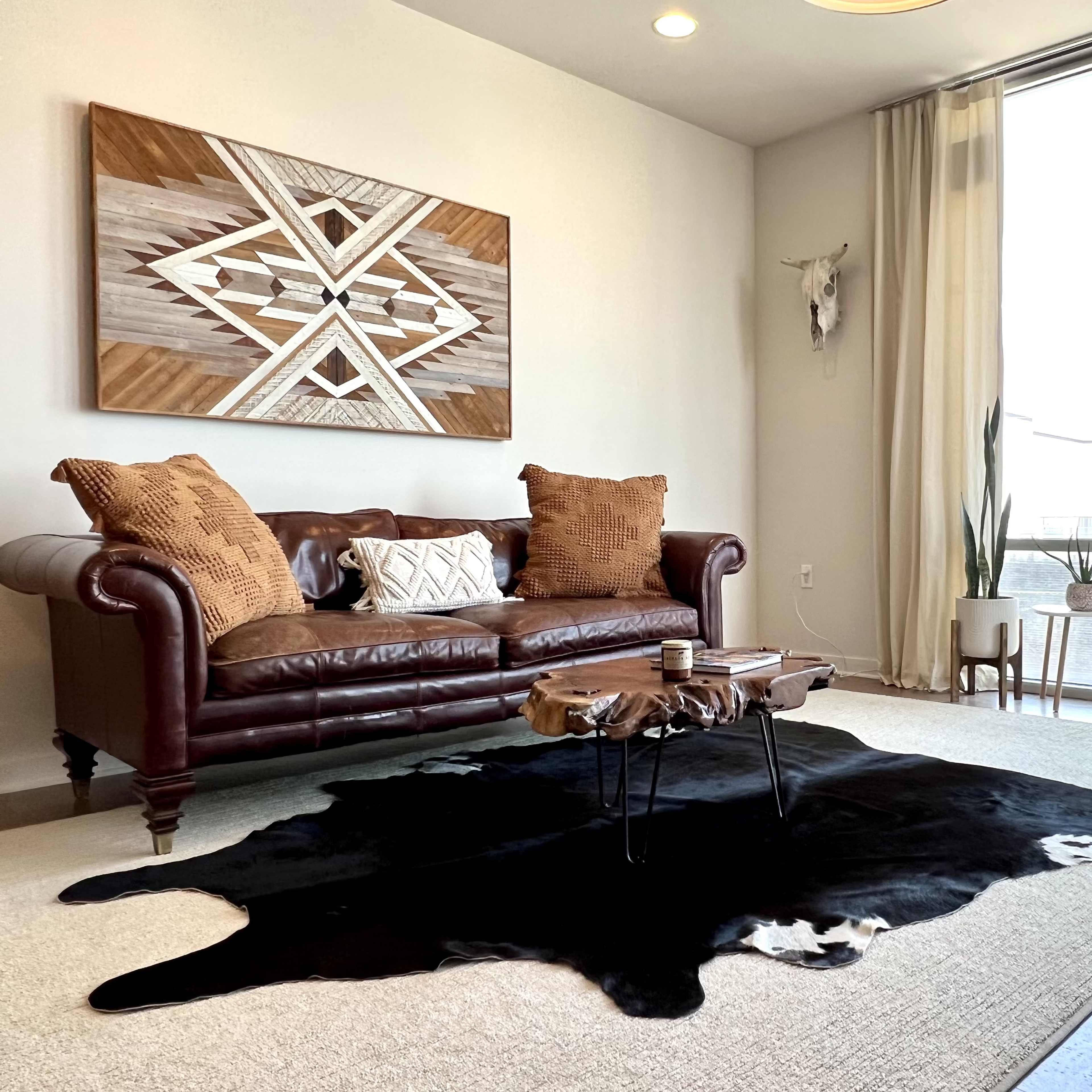 A modern living room featuring a brown leather sofa, a cowhide rug, wooden decor on the wall, and plants in decorative pots beside large windows.