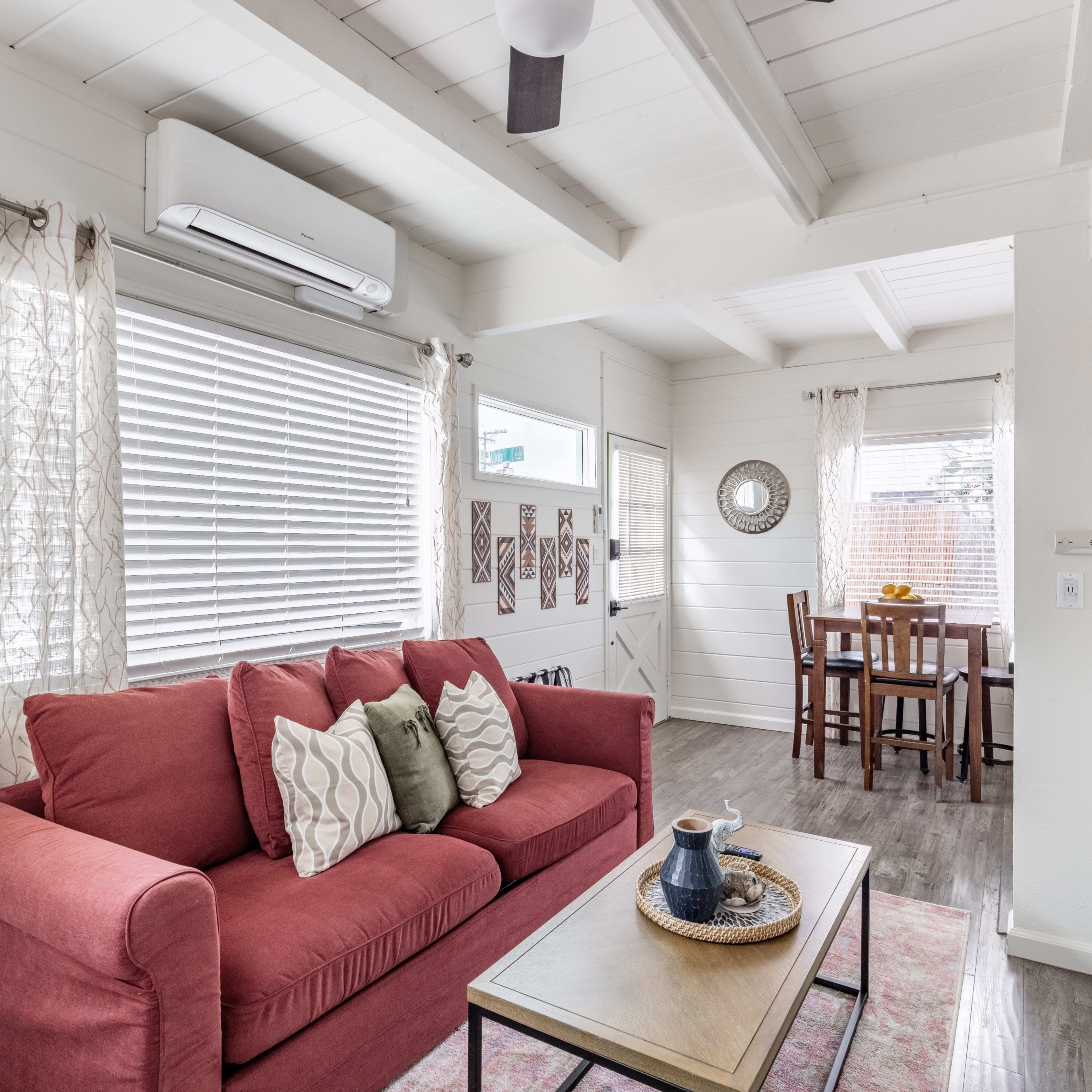 A bright, white interior of a living space with a red sofa, a coffee table, and a dining area with a wooden table and chairs.
