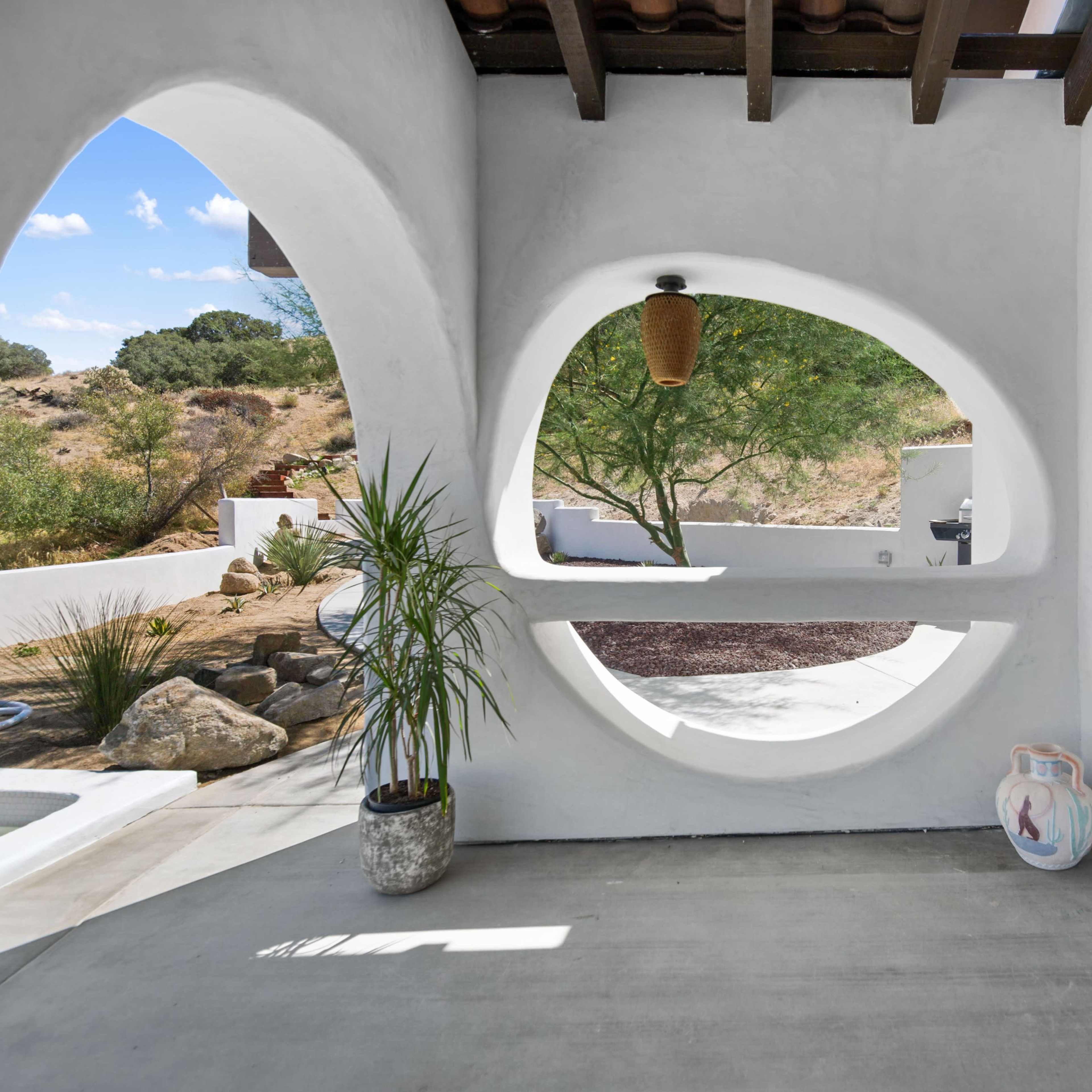A modern architectural space featuring a white stucco wall with circular windows, a concrete floor, and a few plants.