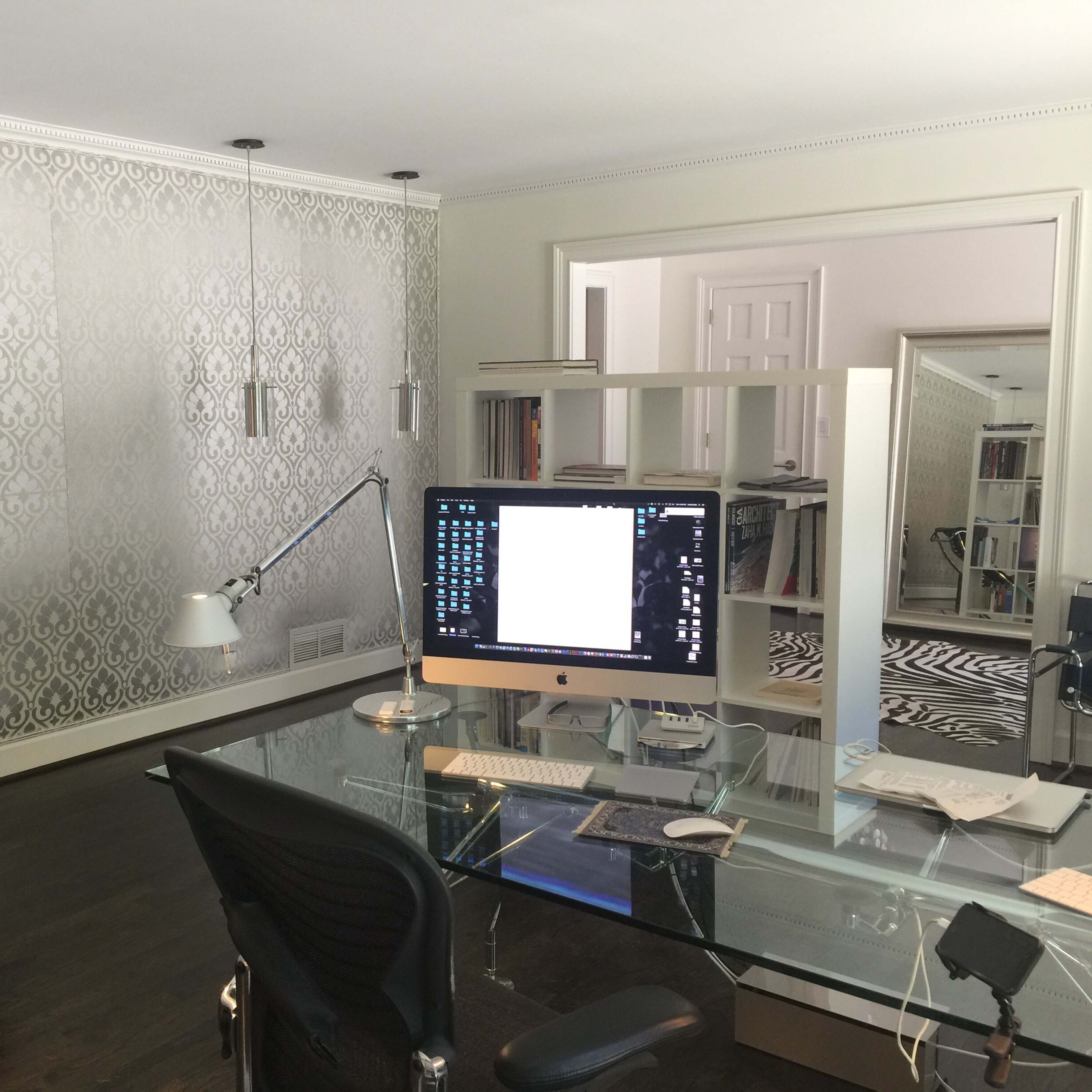 A modern office space featuring a glass desk with an iMac, a black lounge chair, and a patterned wallpaper.