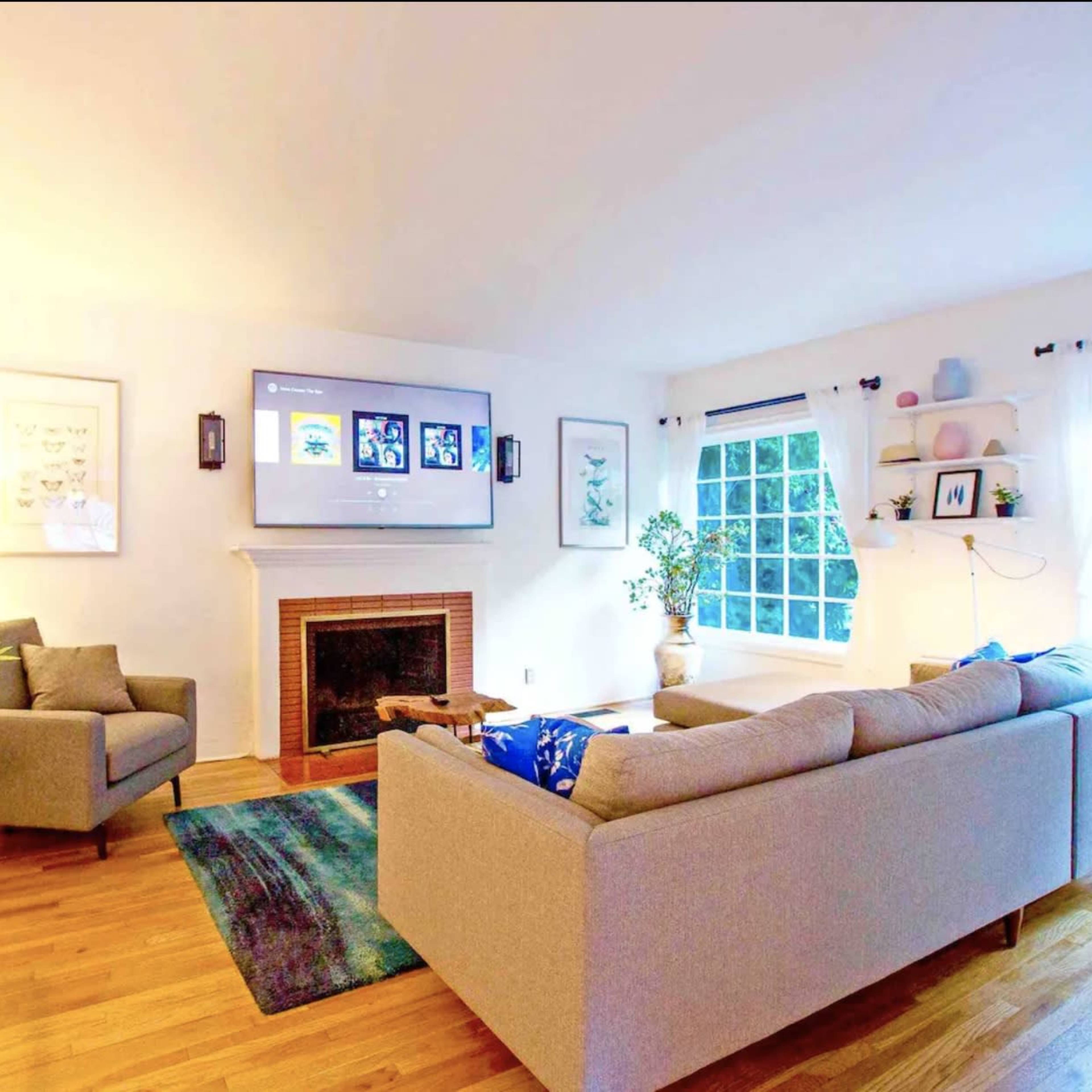 A modern living room with a gray sectional sofa, a fireplace, a TV mounted on the wall, and large windows with white curtains.