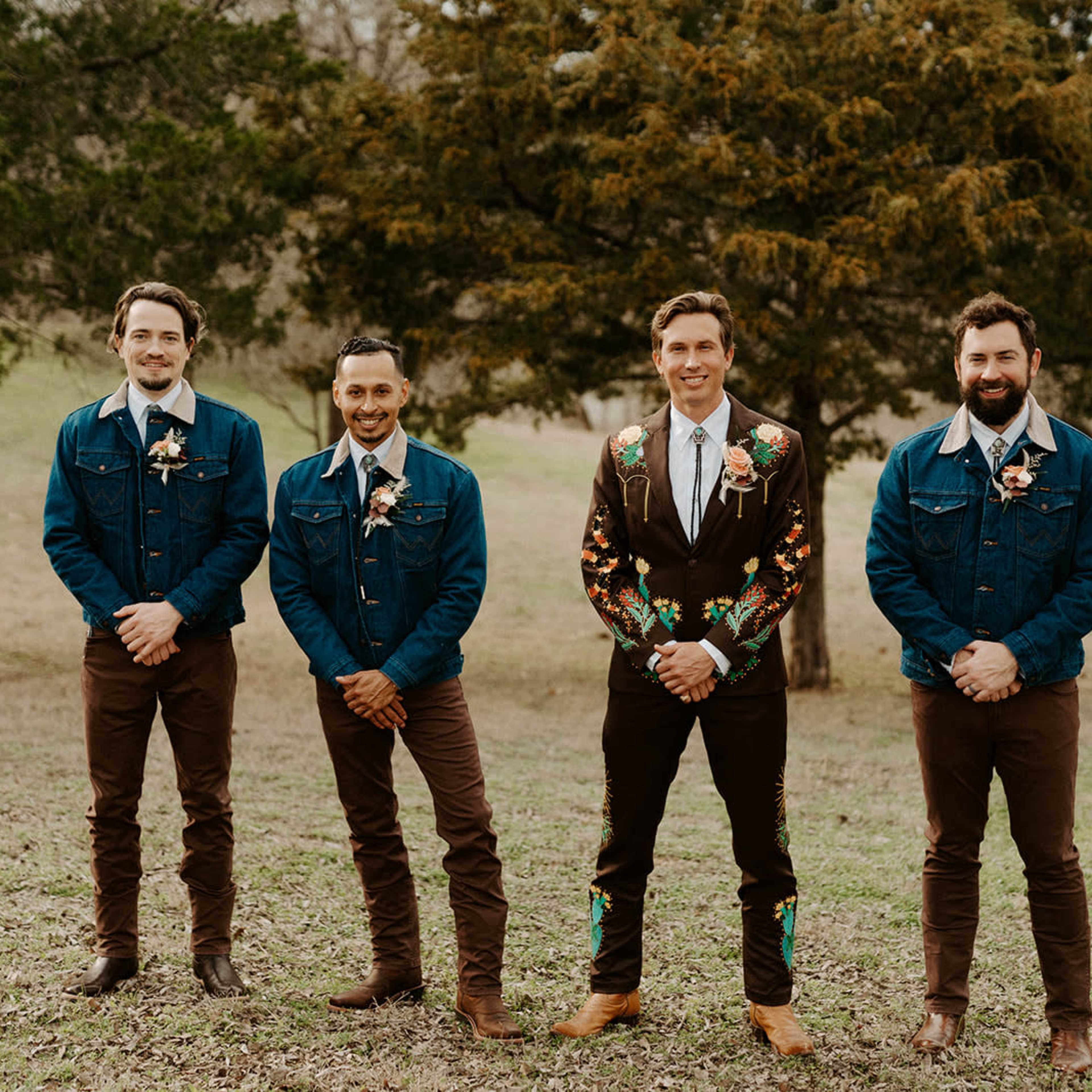 Four men stand in a grassy area, wearing denim jackets or decorated suits with floral designs, while posing for a photo.