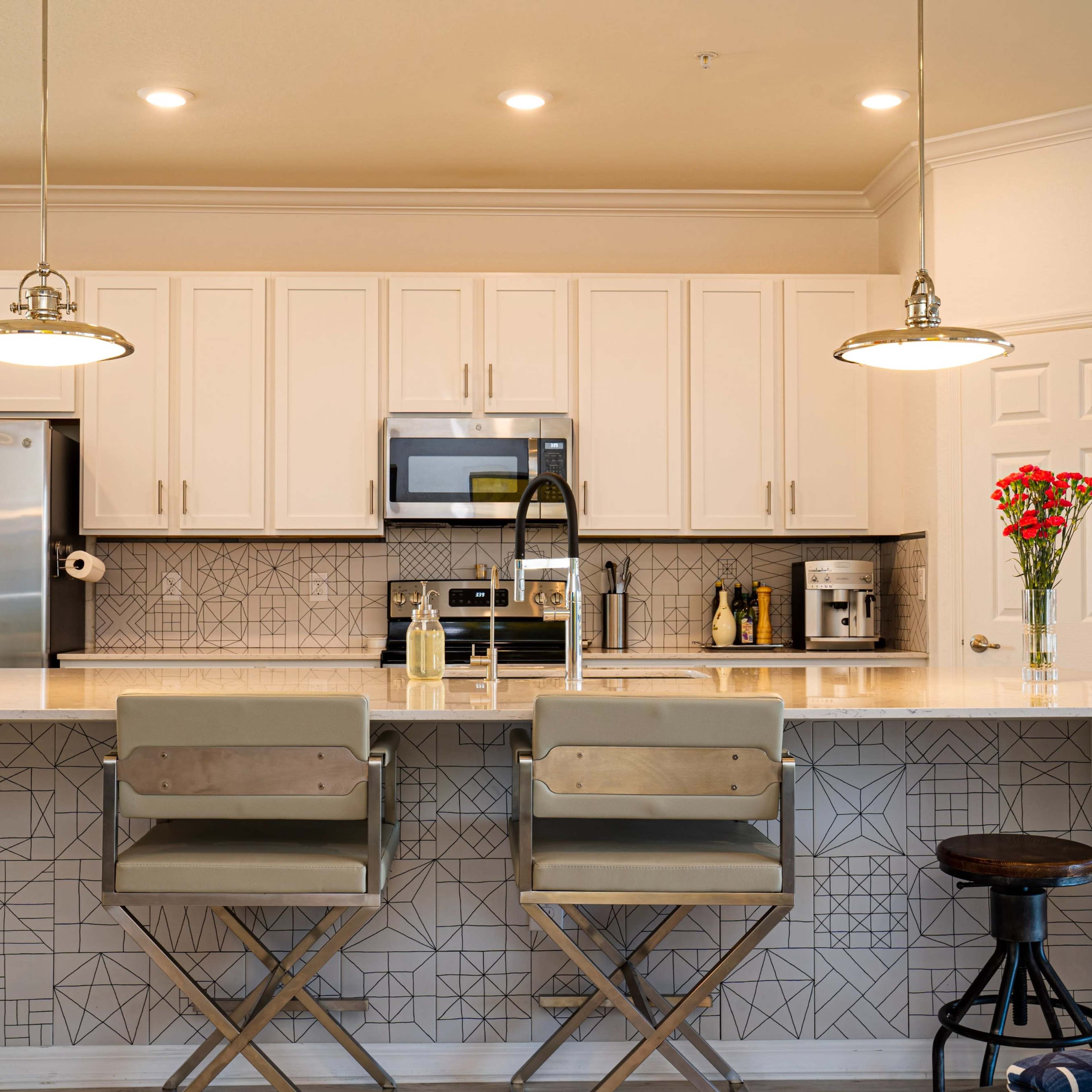 A modern kitchen with white cabinetry, stainless steel appliances, and a central island featuring bar stools and decorative tile work.