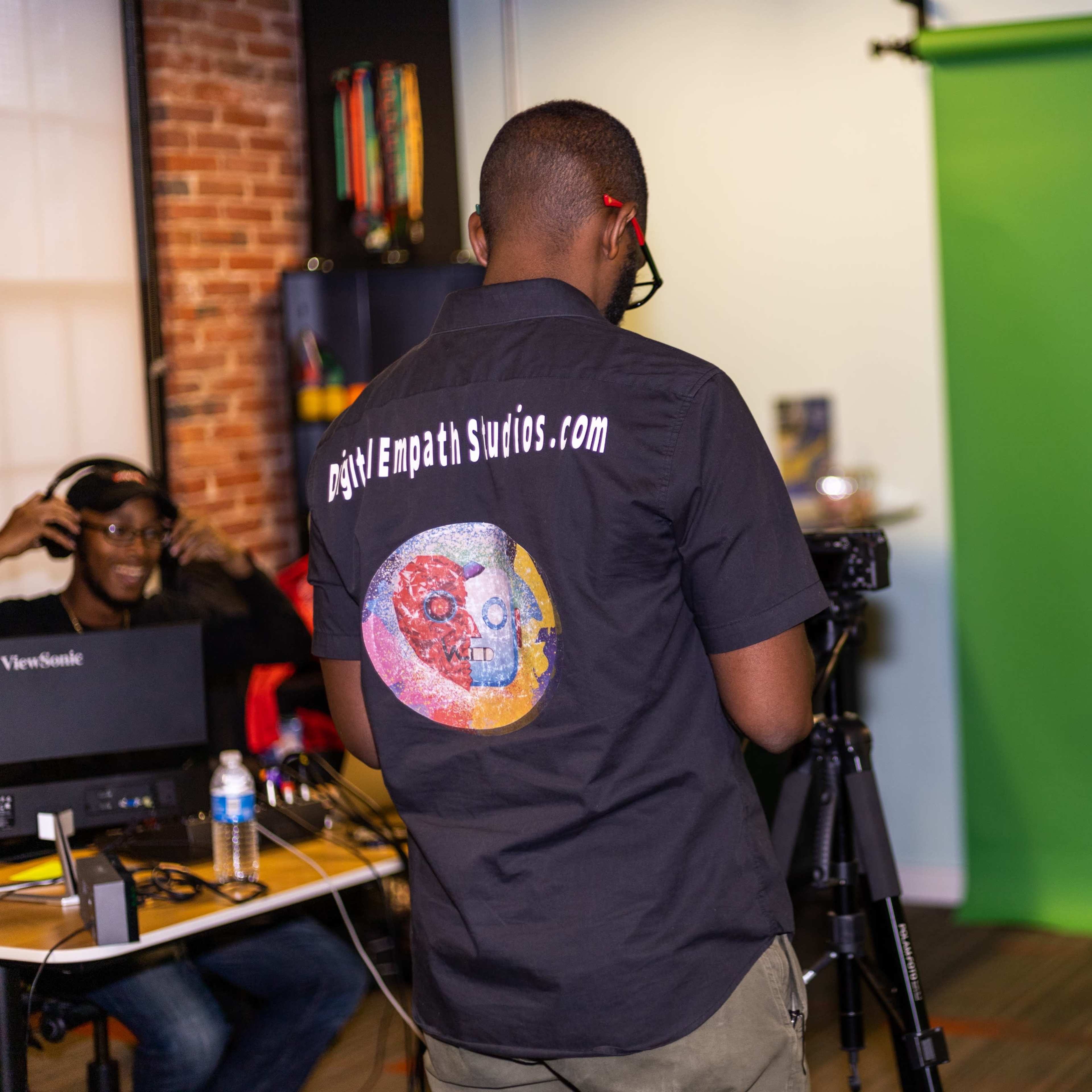 A video production setup features a person in a black shirt with a logo directing a seated individual in front of a green screen, while another person operates equipment nearby.