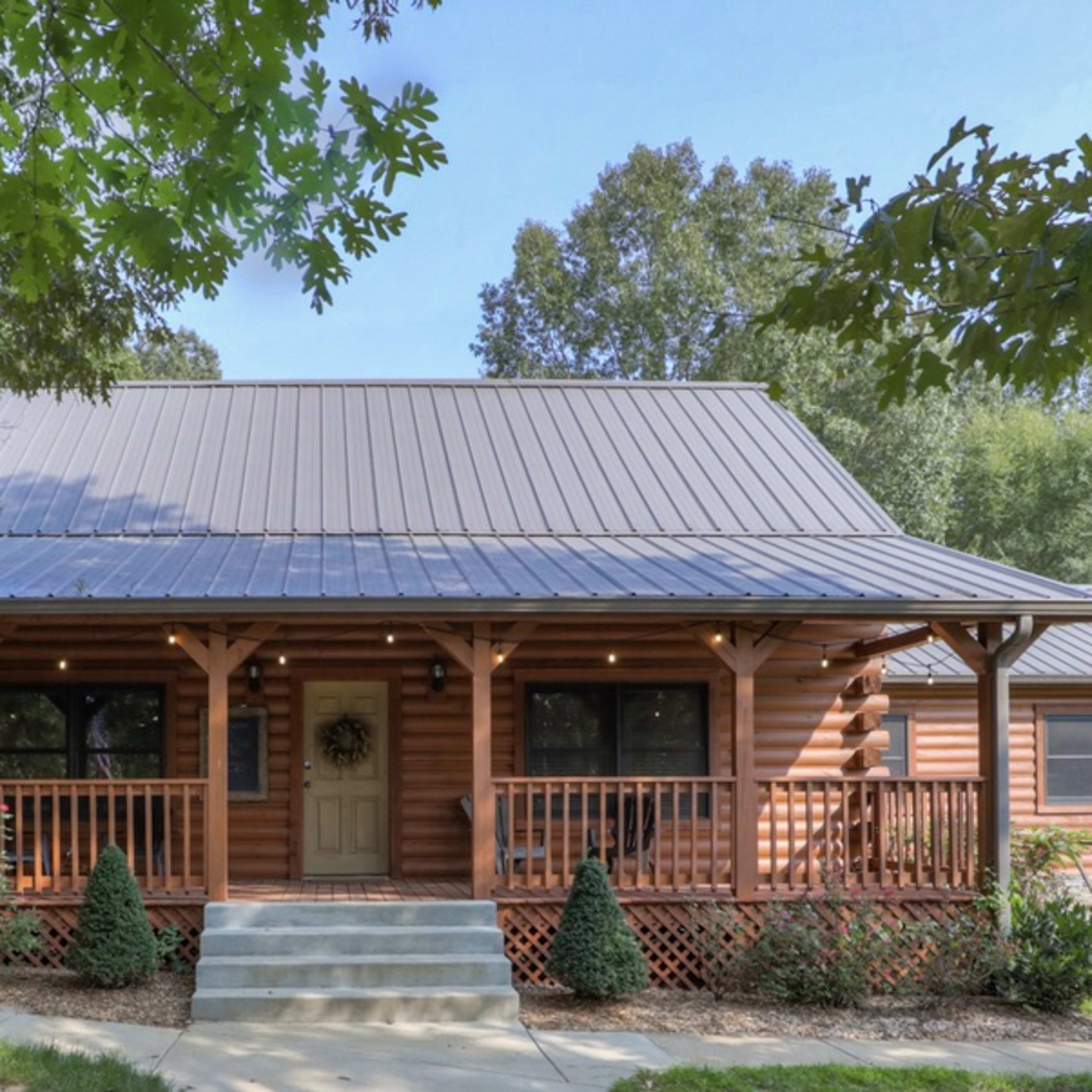 A log cabin-style house with a metal roof, surrounded by trees and featuring a front porch with decorative railing and a flag.