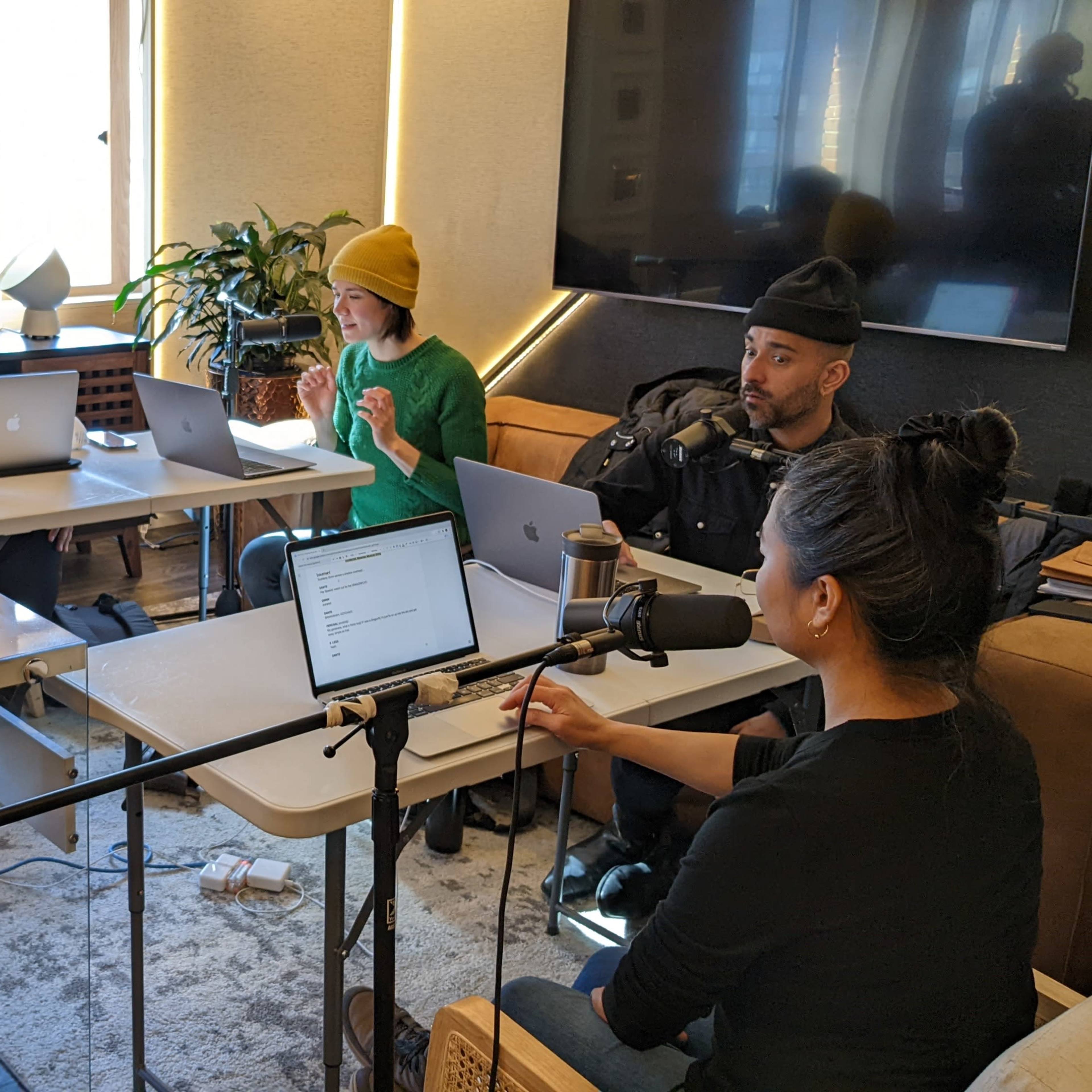 A group of five people are seated around tables with laptops and microphones set up in a modern office space.