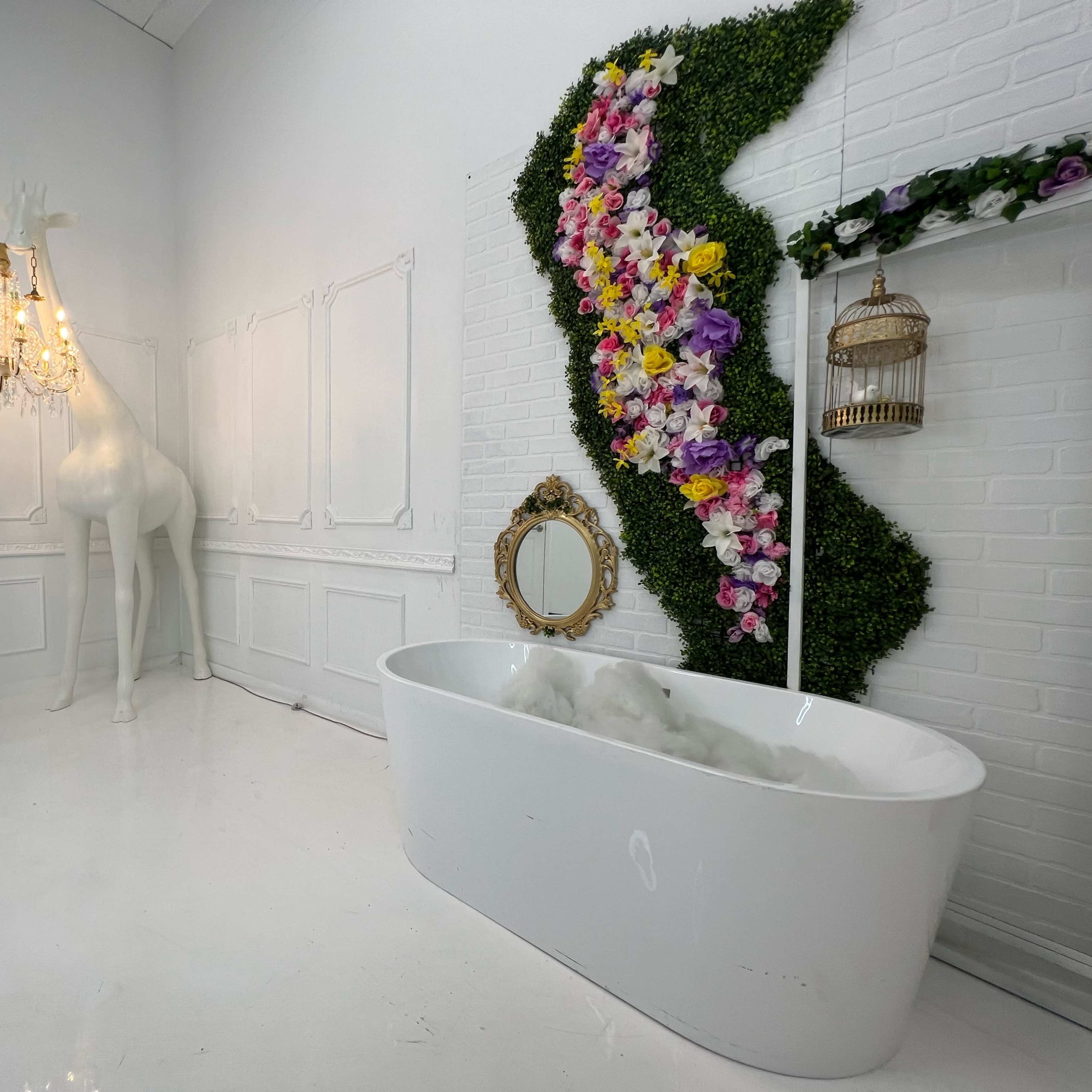 A modern bathroom with a freestanding white bathtub, a decorative wall of flowers and greenery, a large white giraffe sculpture, and a chandelier.