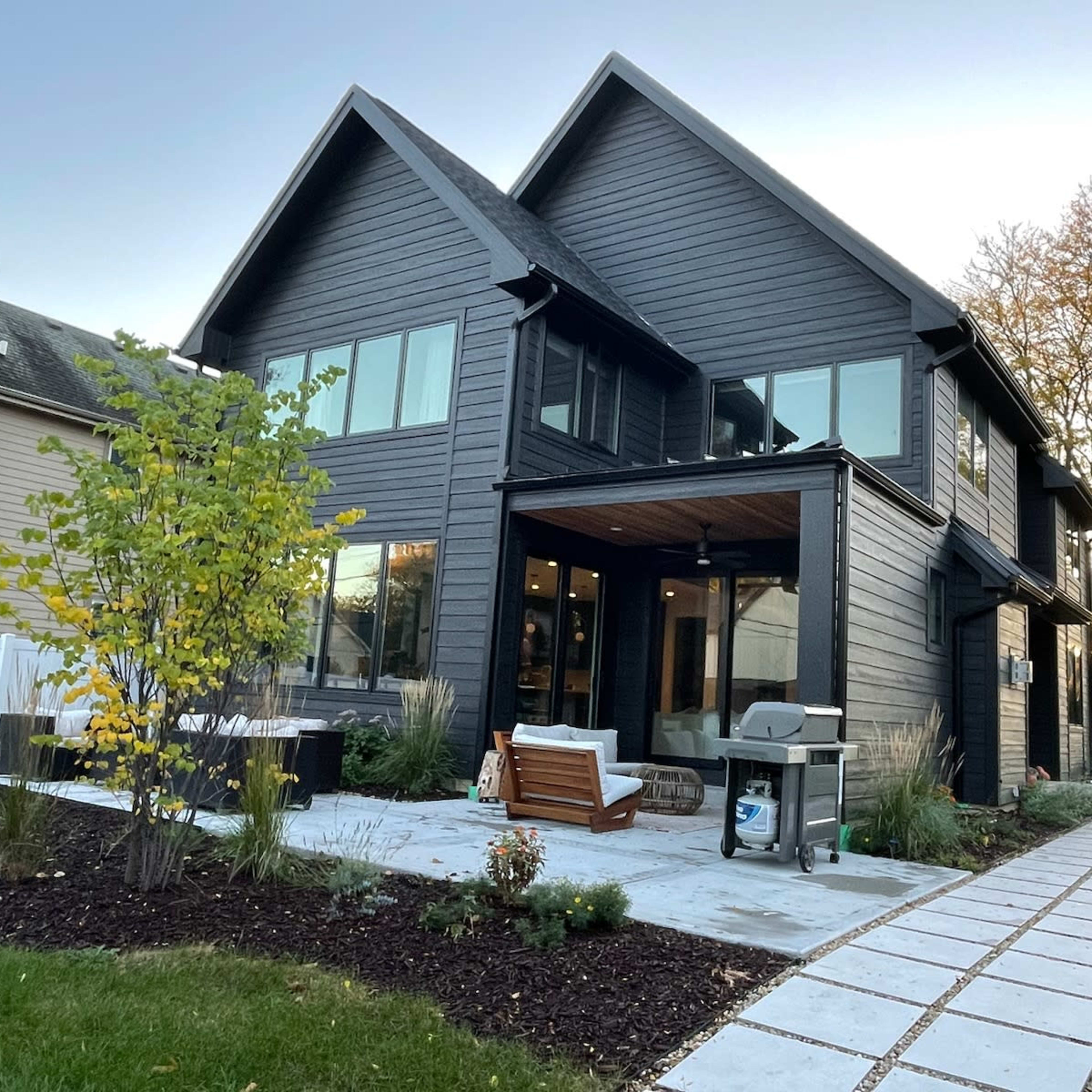 A modern black wooden house with large windows, a covered porch, and a neatly landscaped yard featuring a pathway and plants.