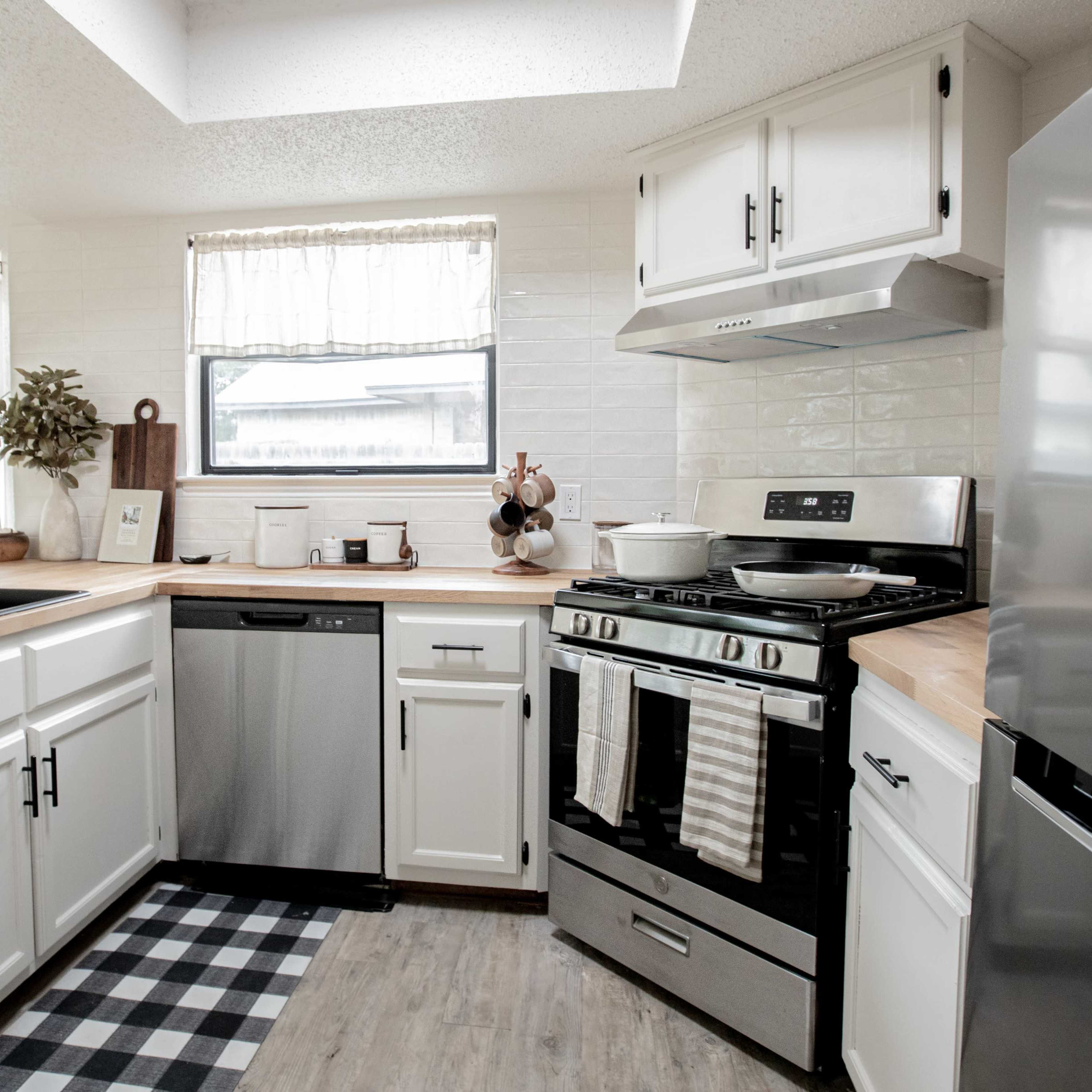 The kitchen features white cabinets, a stainless steel sink, a dishwasher, and a gas stove on a wood countertop, with large windows providing natural light.