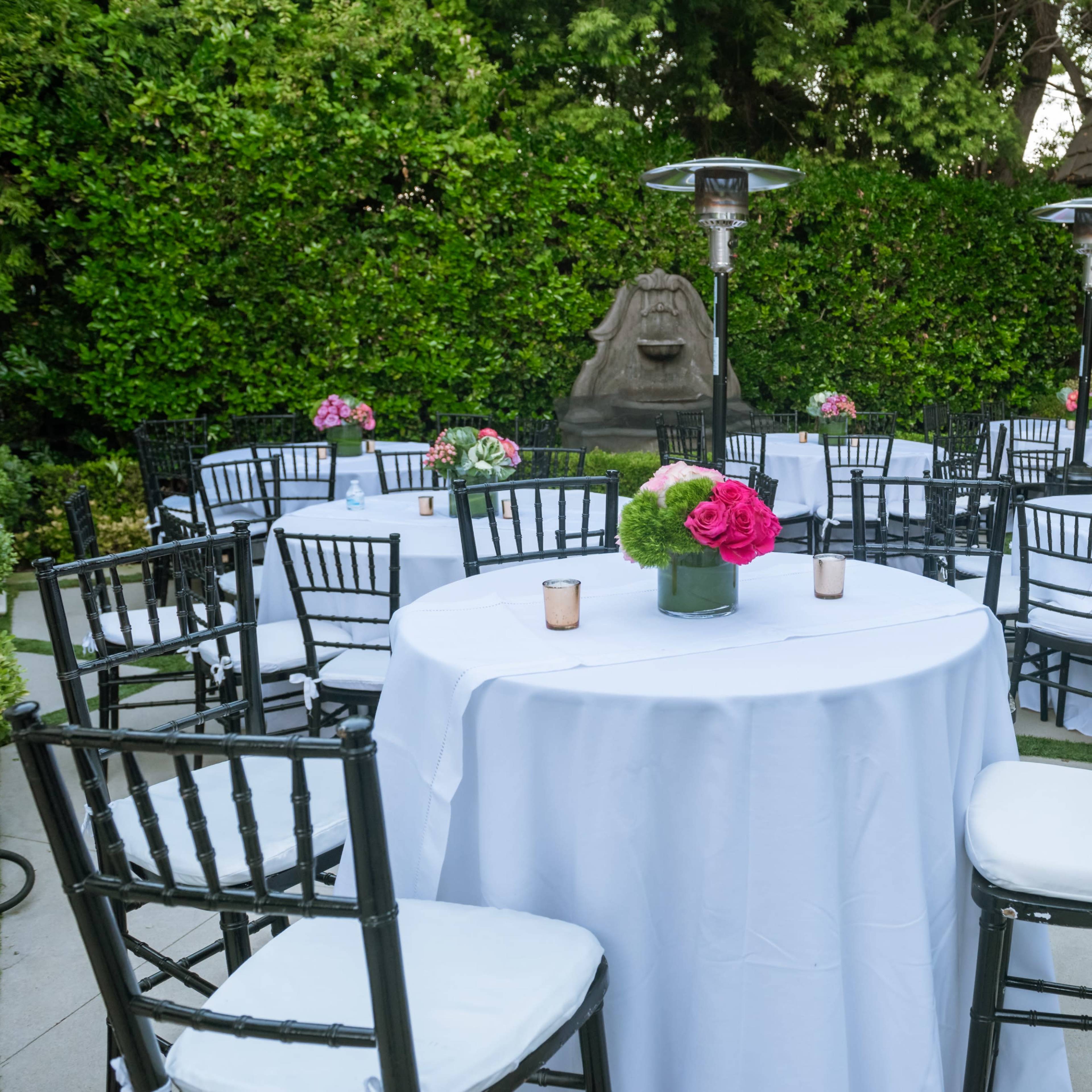 A garden setting features round tables covered with white tablecloths, surrounded by black chairs, and adorned with centerpieces of flowers.