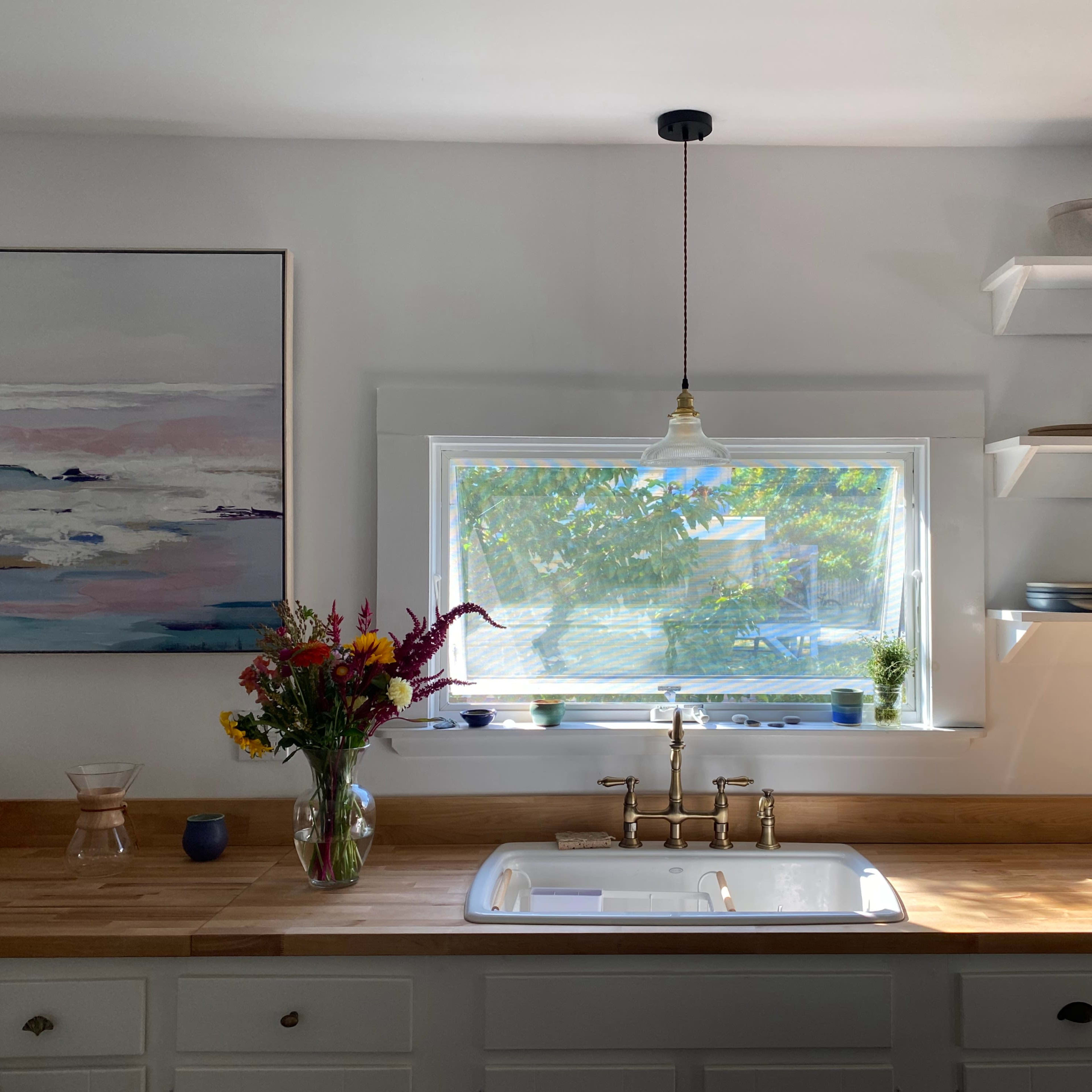A kitchen features a window above a sink, surrounded by shelves with dishes and a bouquet of flowers on the countertop.