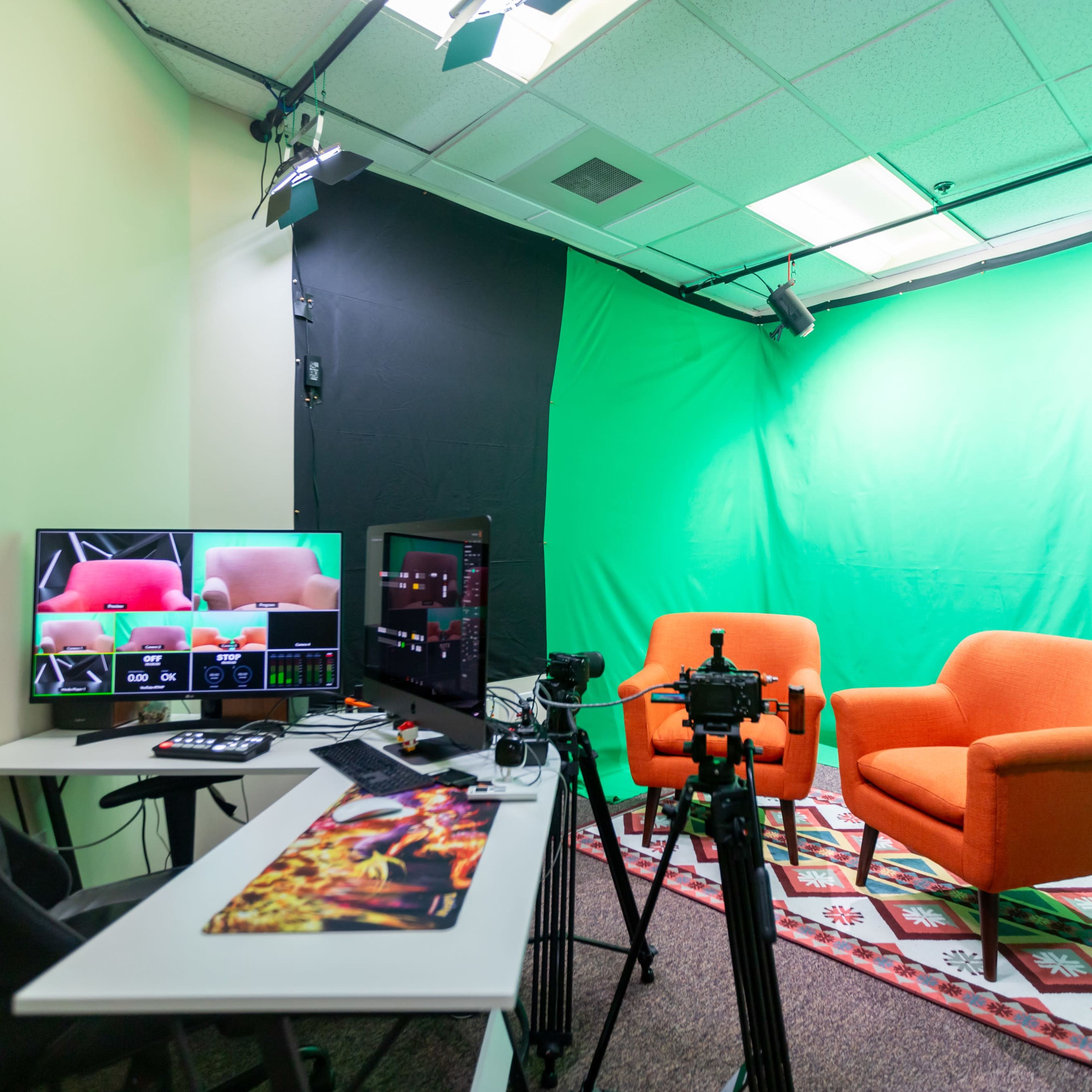 A recording studio featuring two orange chairs on a patterned rug, a computer setup with multiple screens, and a green screen backdrop.