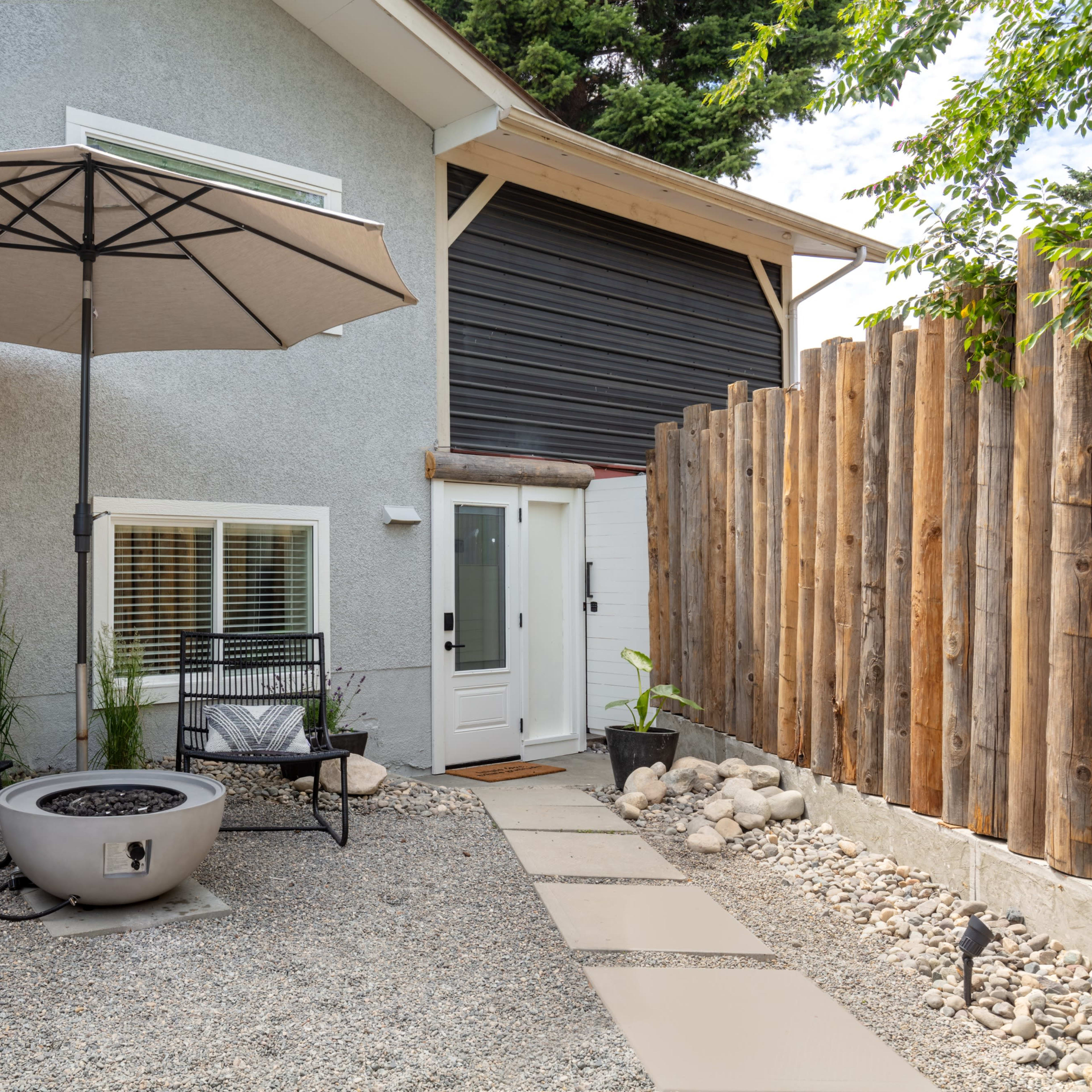 A backyard patio area with two black chairs, a round fire pit, a large umbrella, and a gravel path leading to a house with a wooden fence.