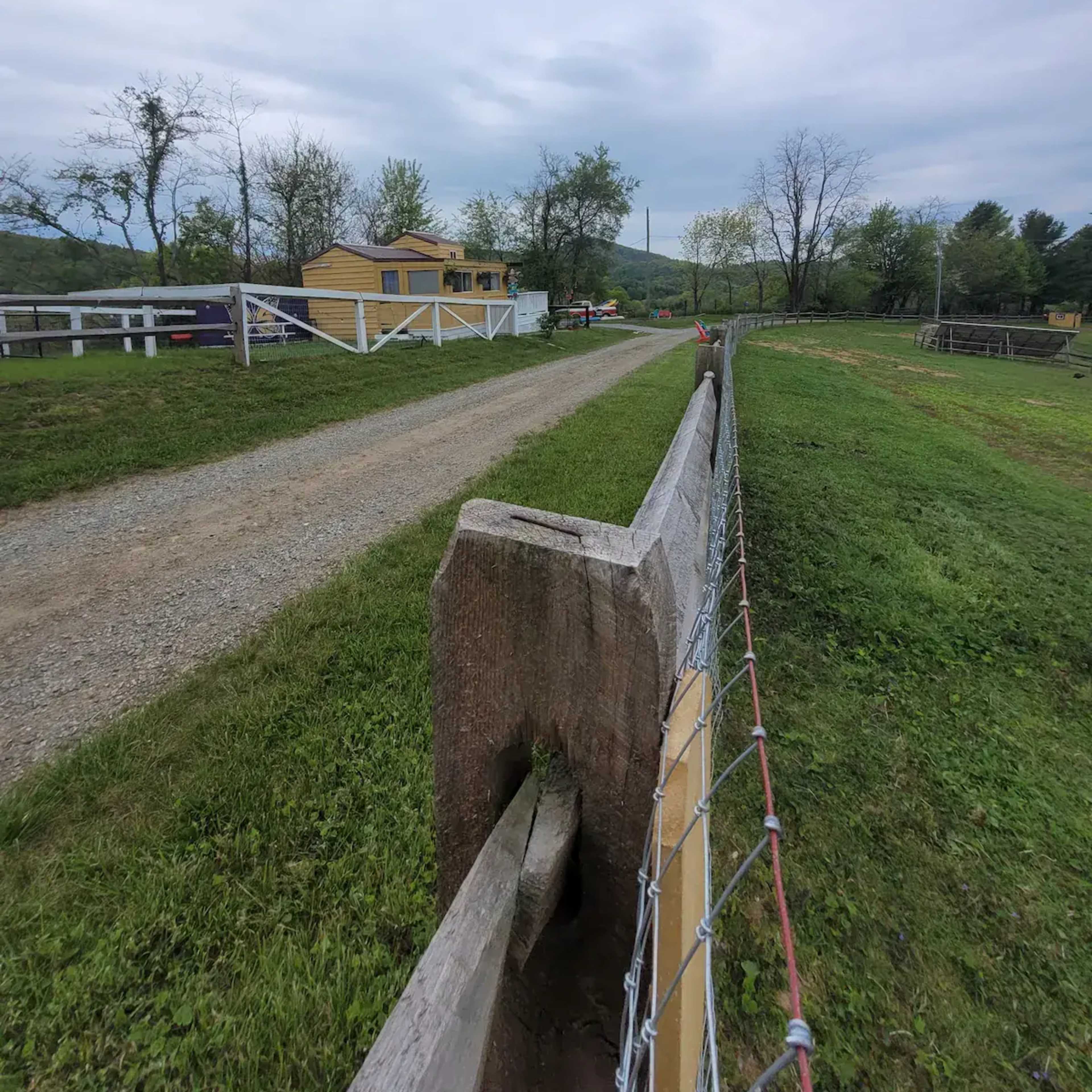 A gravel road leads towards a yellow house surrounded by a grassy landscape and a wire fence.