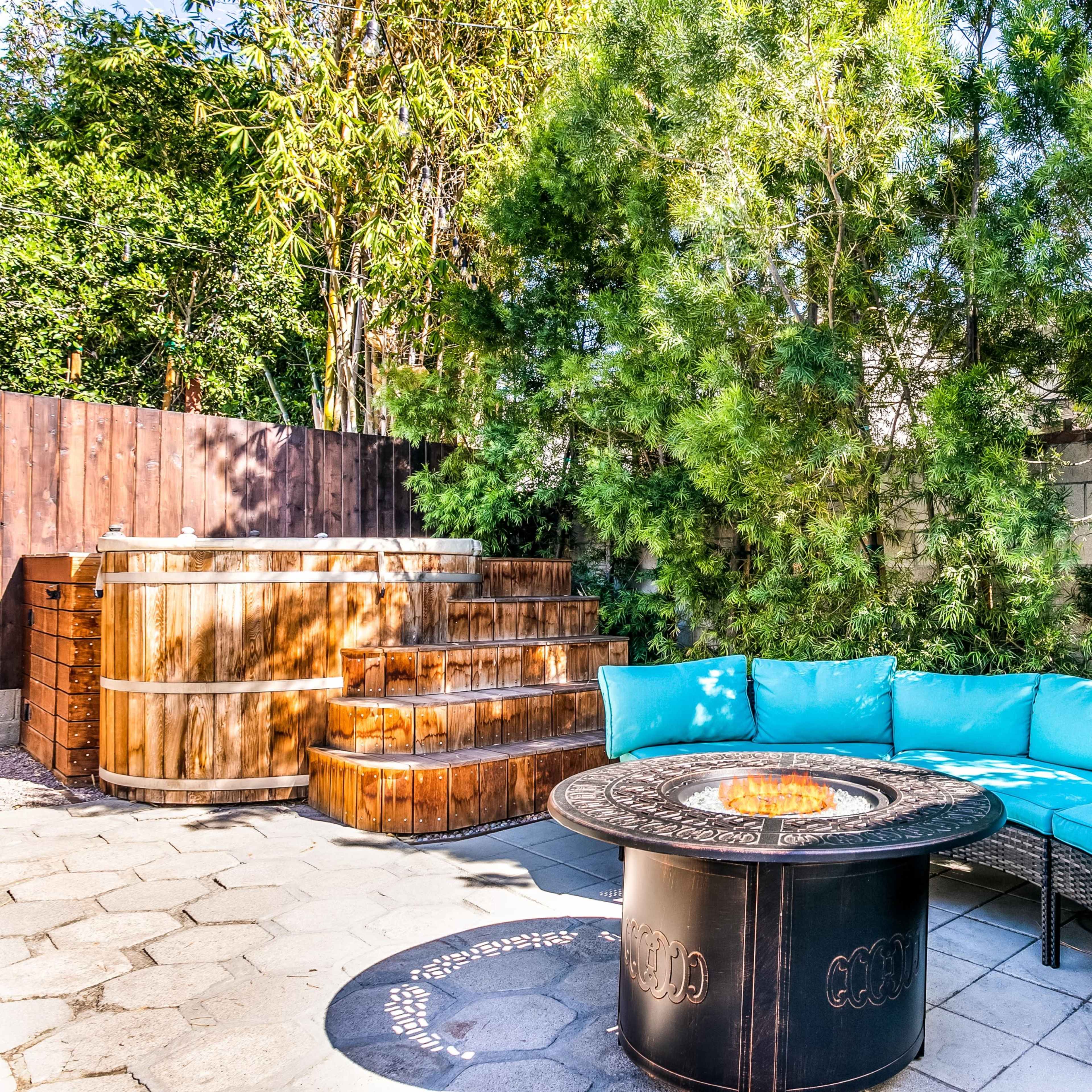 A backyard patio with a blue sectional sofa surrounding a circular fire pit, wooden hot tubs on a raised platform, and lush greenery in the background.