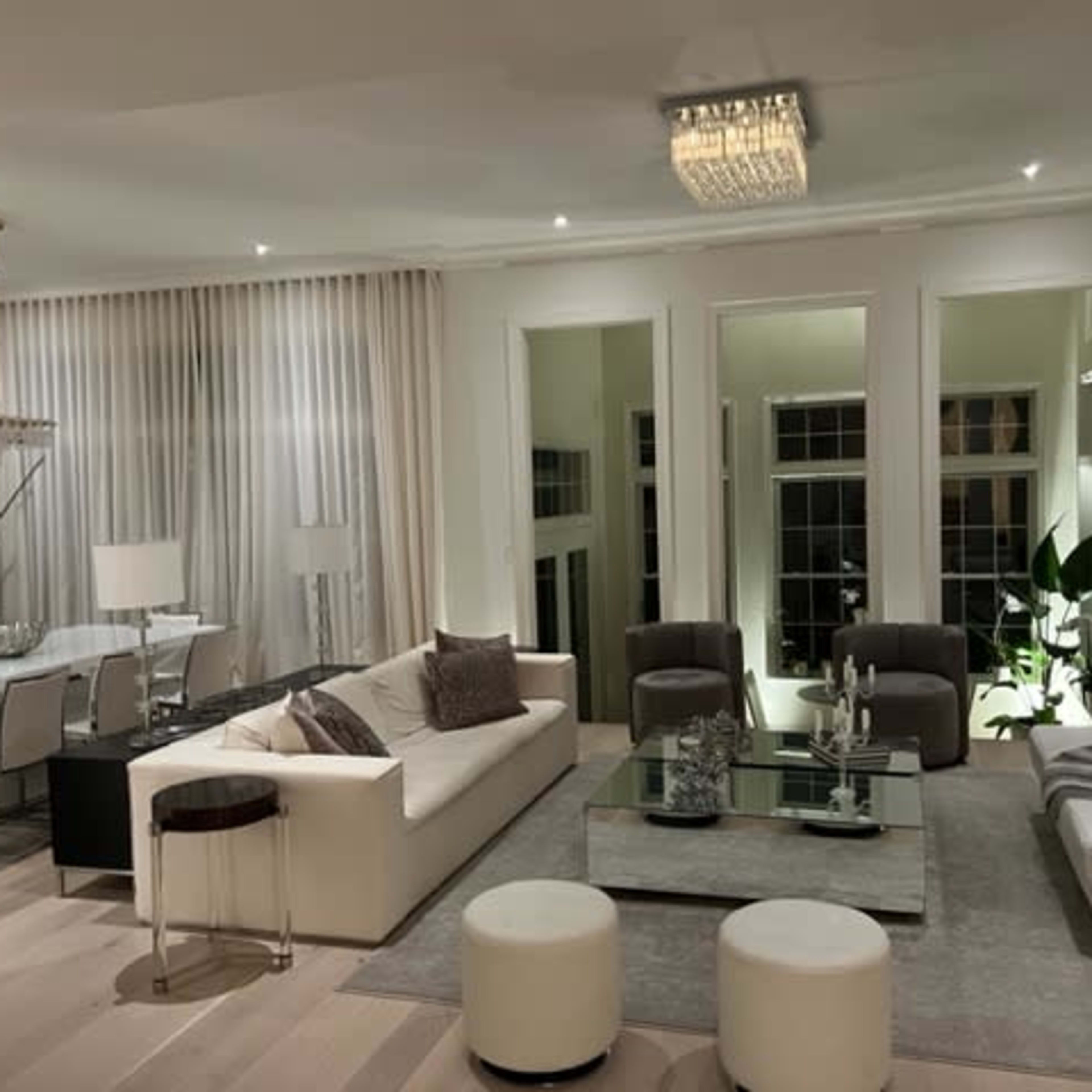 A modern living room with a white sofa, two brown chairs, a glass coffee table, and large windows, complemented by light-colored walls and elegant lighting.